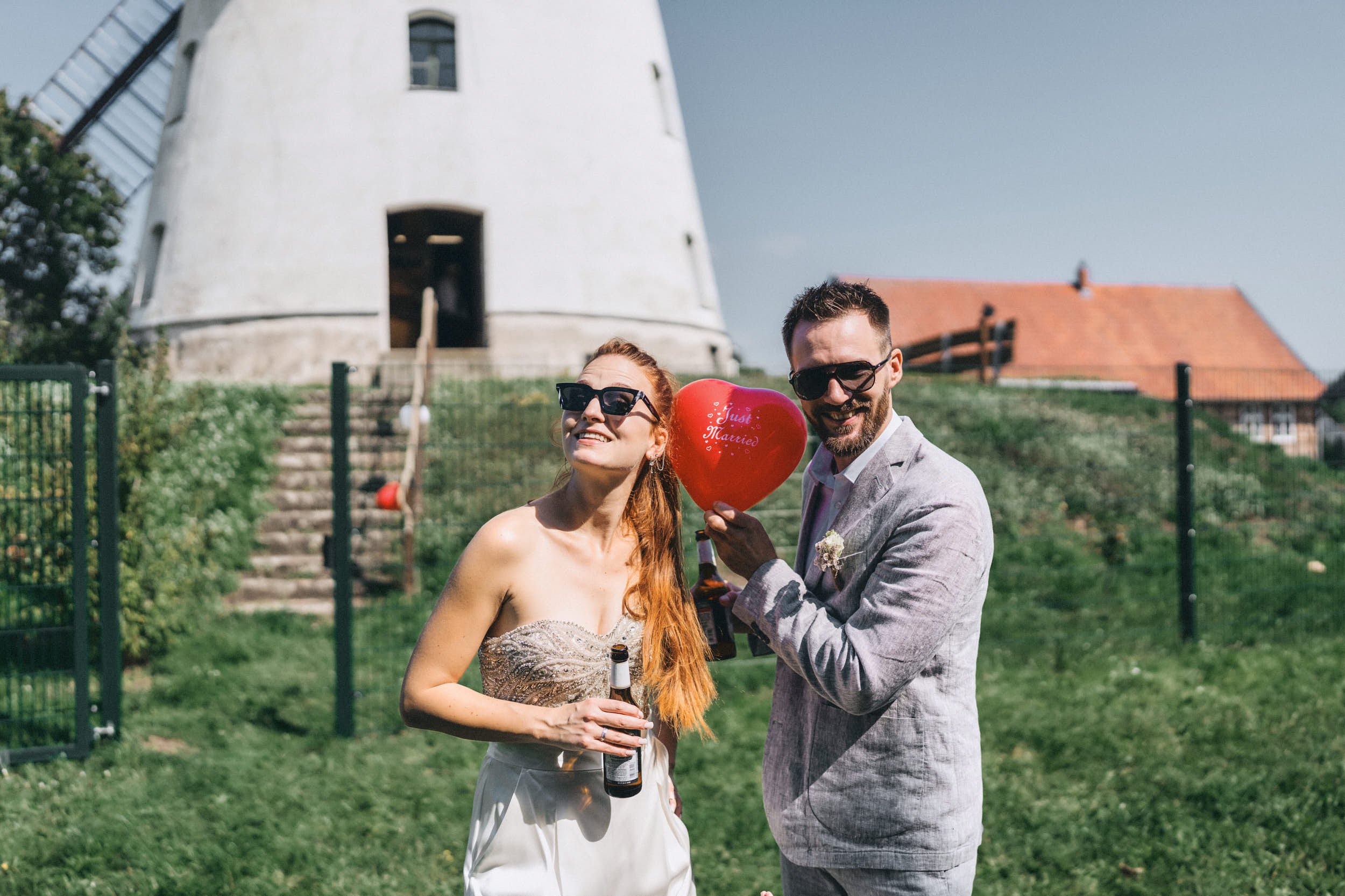 Ein lachendes Paar bei einer Hochzeit im Garten, die Frau hat rote Haare und trägt ein Brautkleid, der Mann trägt einen grauen Anzug, beide tragen Sonnenbrillen, er hält ein Herz mit der Aufschrift "Just Married" und eine Bierflasche, im Hintergrund 