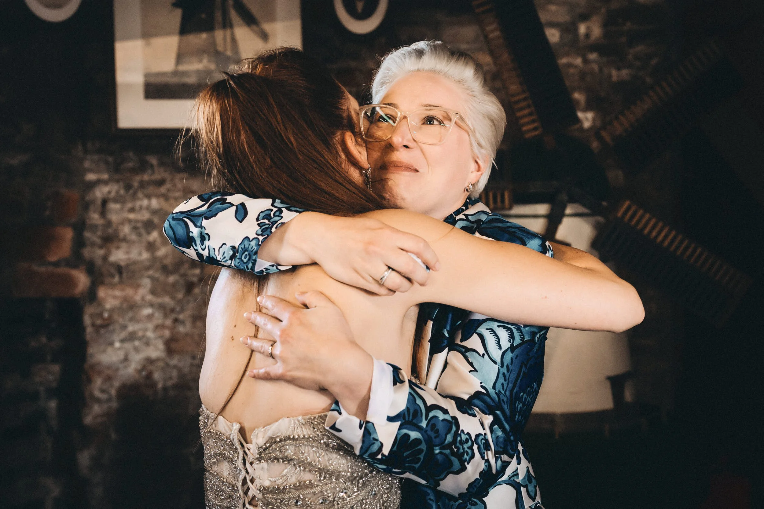 Zwei Frauen umarmen sich herzlich, eine mit weißen Haaren und Brille, die andere mit braunen Haaren, in einem gemütlichen Raum mit dunklen Wänden und einem Wandbild im Hintergrund.