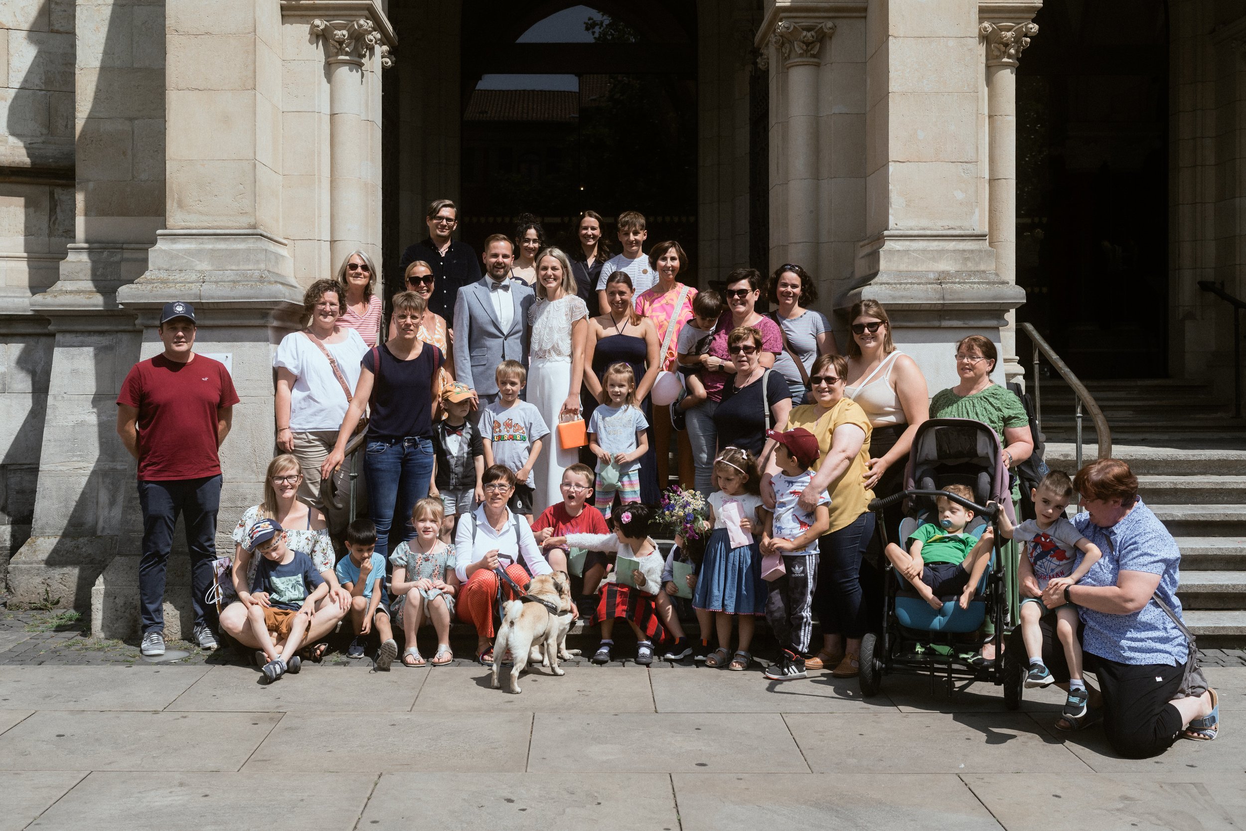 Gruppe von Menschen, inklusive Kinder, Frauen und Männern, auf den Stufen vor einem historischen Gebäude mit Säulen. Manche Personen tragen Sonnenbrillen und haben einen Hund dabei.