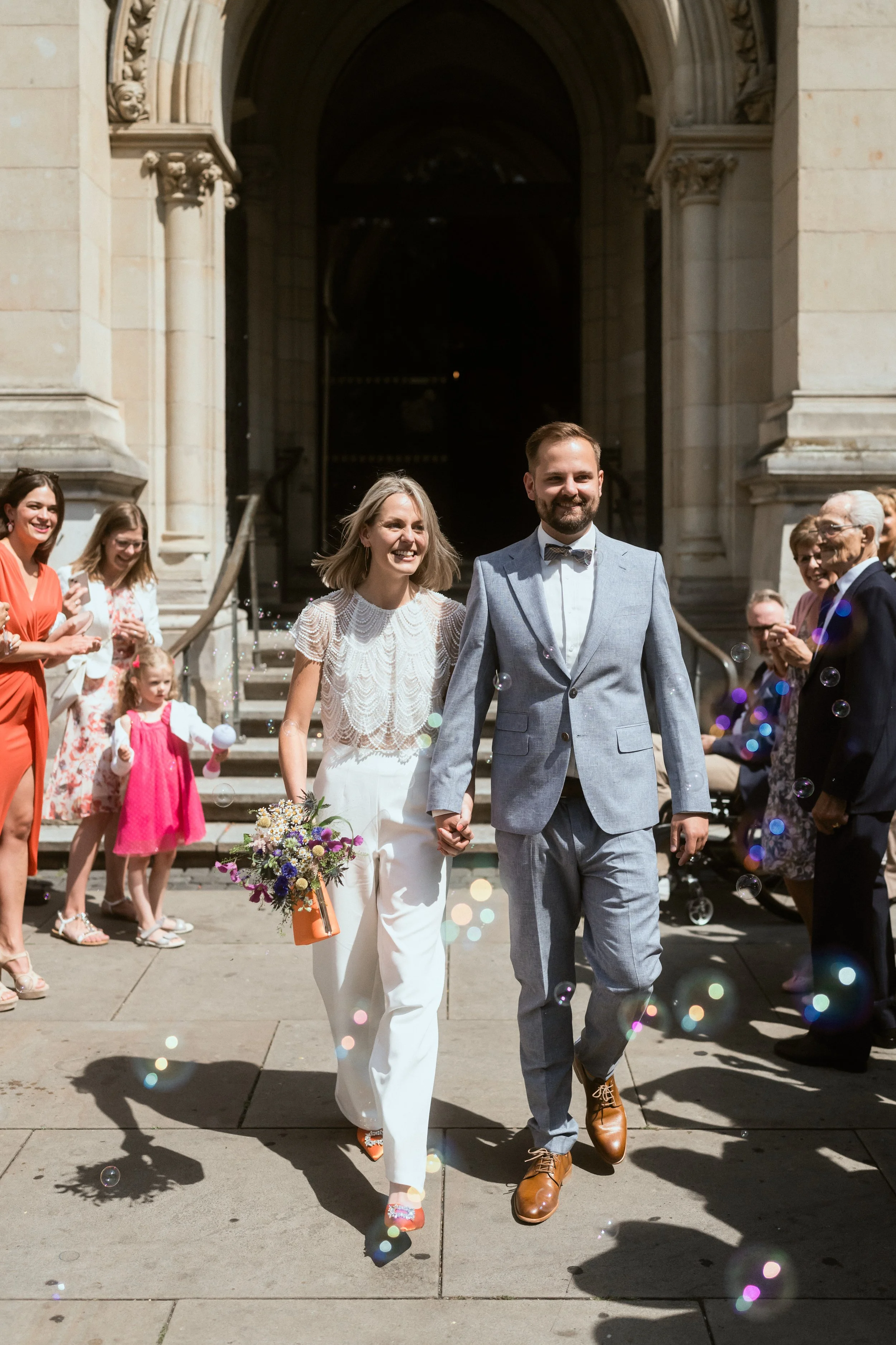 Ein Hochzeitsfoto zeigt ein lachendes Brautpaar, das Hand in Hand vor einer Kirche ausgeht, umgeben von Gästen, die feiern, mit Seifenblasen in der Luft an einem sonnigen Tag.