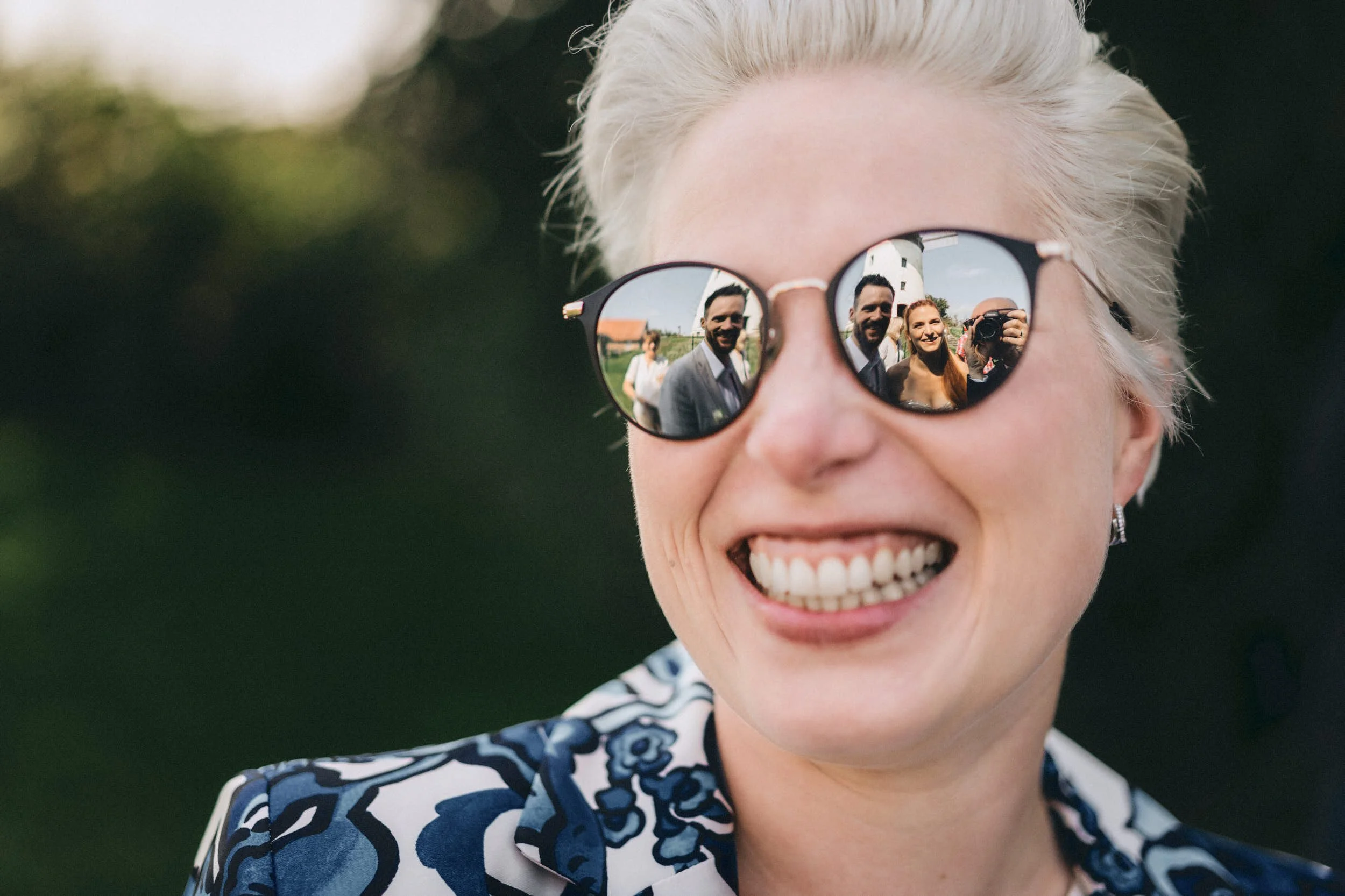 Eine lachende Frau mit kurzen, platinblonden Haaren trägt runde Sonnenbrillen, die die Spiegelung eines Mannes und einer Frau zeigen, die sich gegenseitig fotografieren, im Freien bei Sonnenlicht.