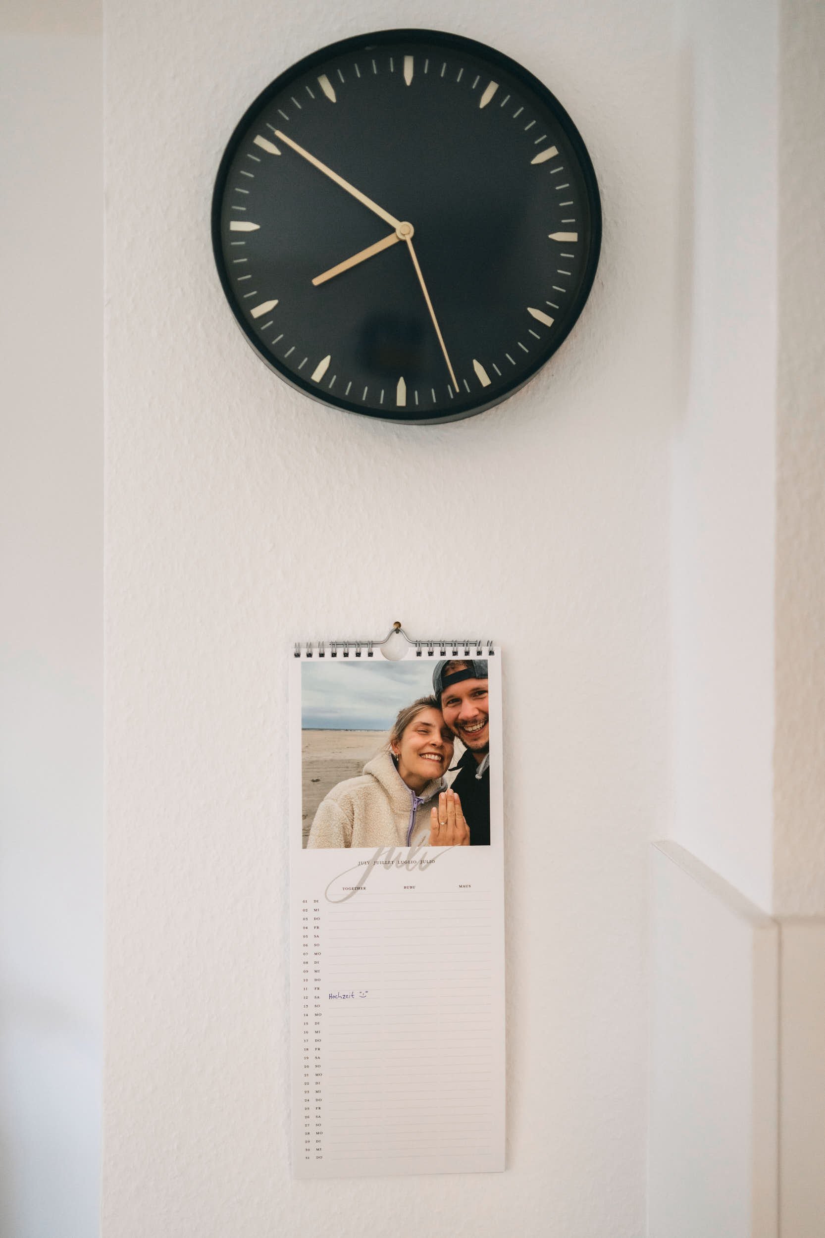 Eine Uhr an einer weißen Wand zeigt ungefähr 14:53. Darunter hängt ein Kalender, der ein Foto eines Paares zeigt, das bei einem besonderen Anlass lächelnd zusammensteht.