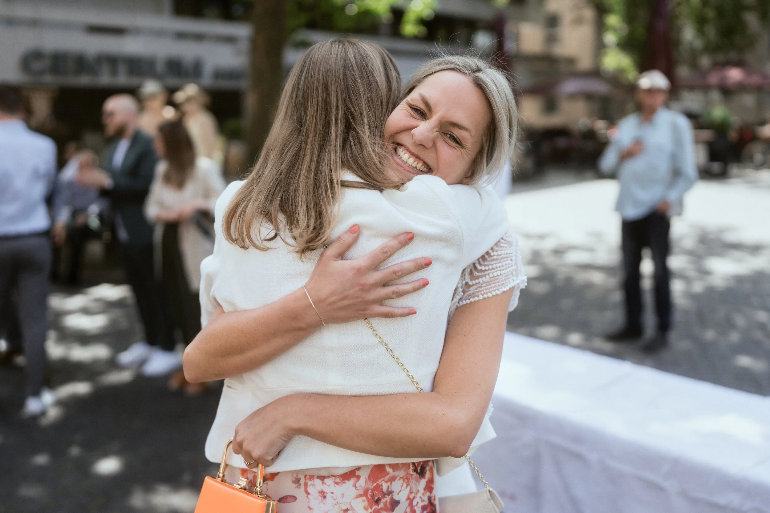 Zwei Frauen umarmen sich freundlich im Freien bei einem gesellschaftlichen Event, im Hintergrund sind weitere Personen zu sehen.