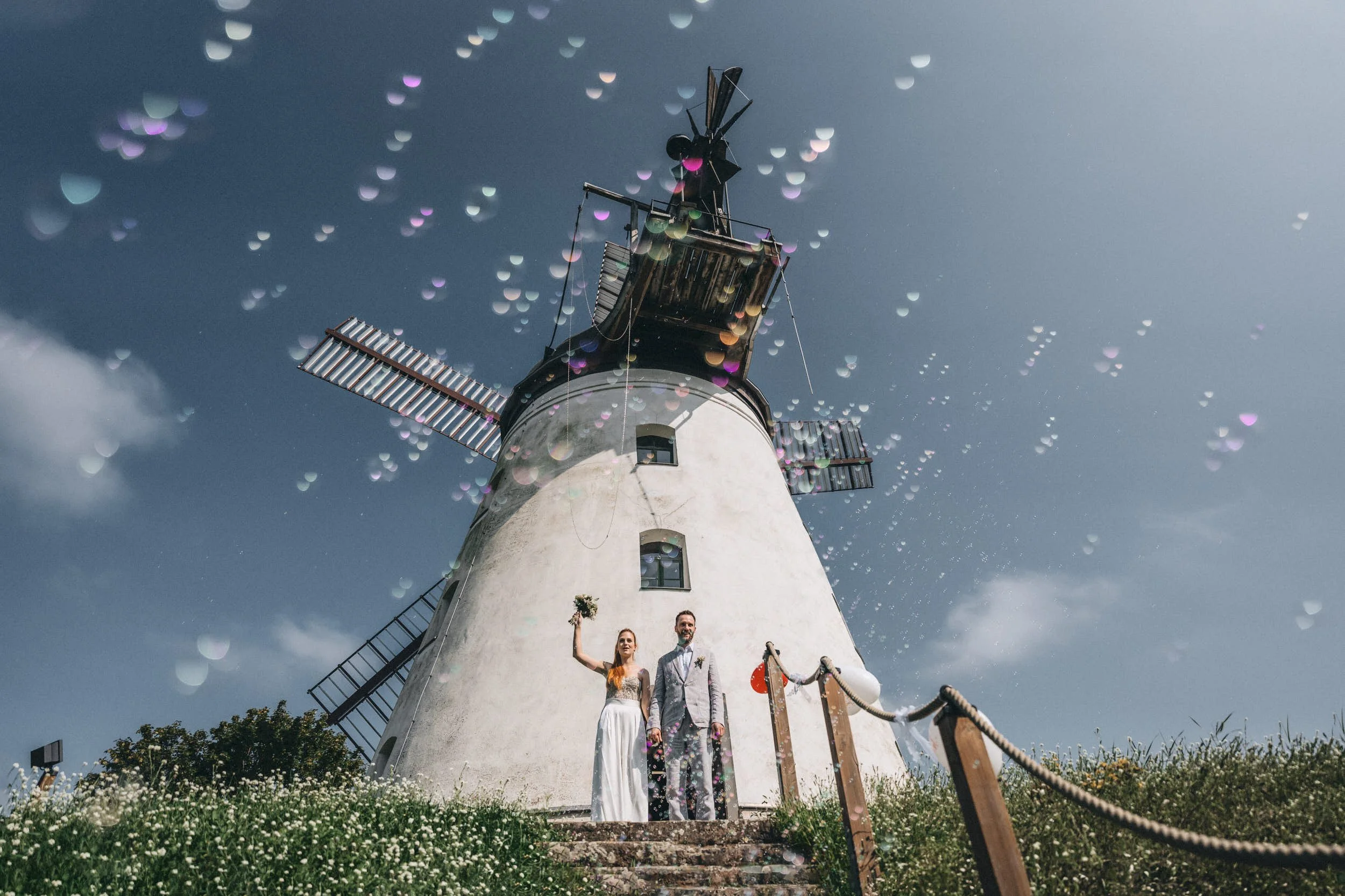 Auf dem Bild sind zwei Personen, ein Mann und eine Frau, die vor einer weißen Windmühle stehen. Der Himmel ist bewölkt, und es gibt bunte Lichtflares, die den Himmel überziehen. Die Windmühle hat vier Flügel und befindet sich in einer grünen Landscha
