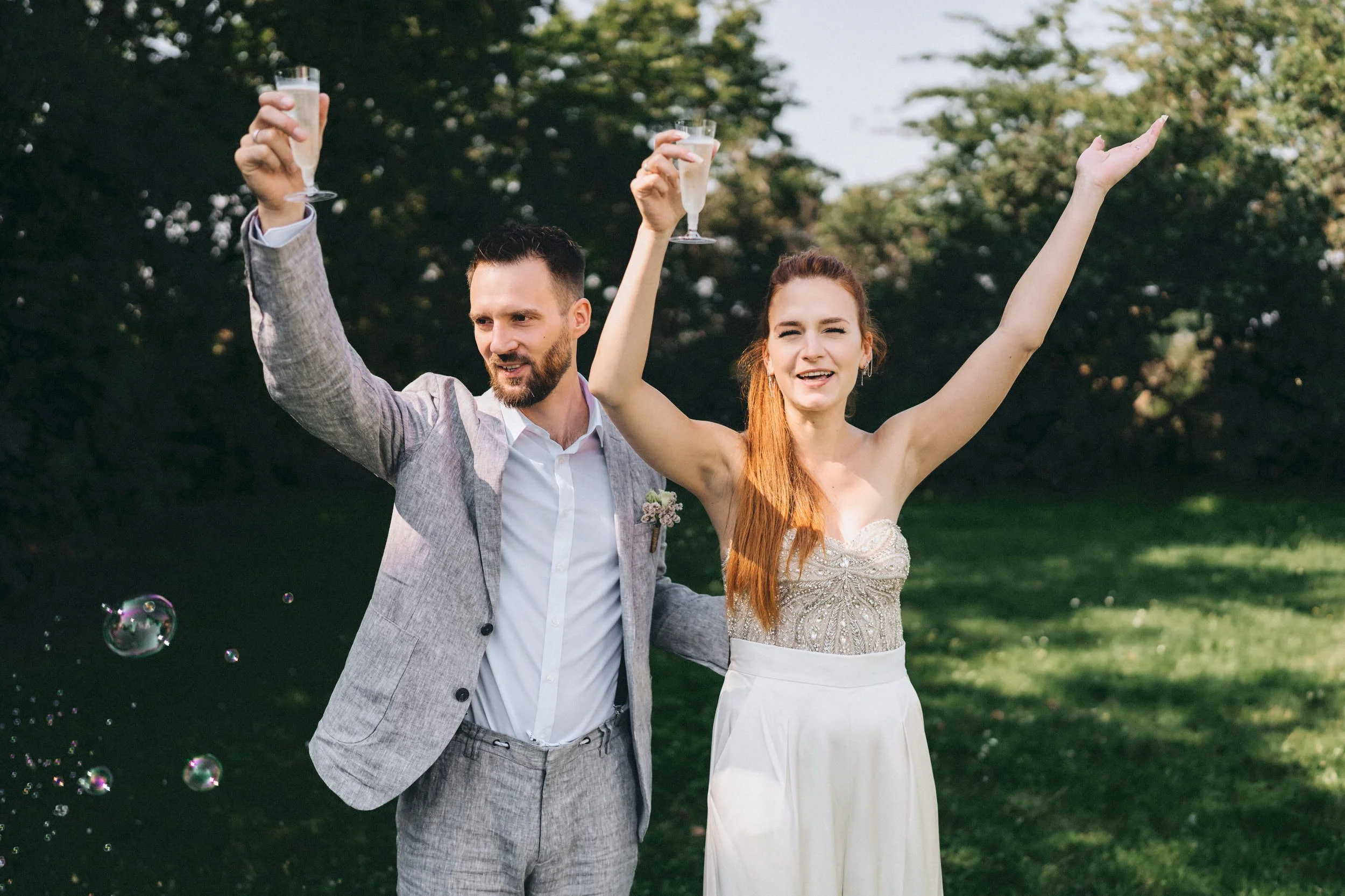 Ein glückliches Paar bei einer Hochzeit im Freien, Anstoßen mit Champagner, im Hintergrund Bäume.