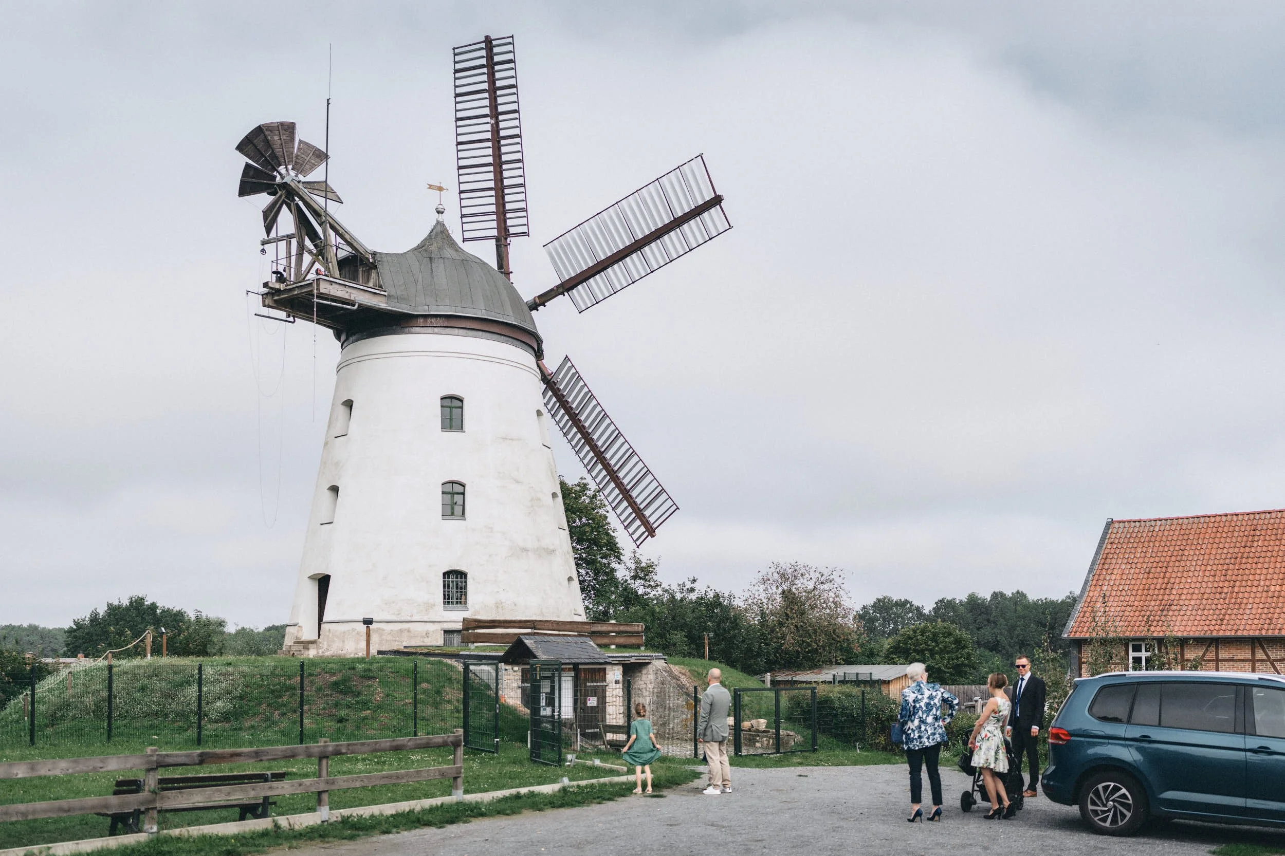 Ein weißer, historischer Windmühle mit vier Flügeln steht im Freien. Im Vordergrund befinden sich mehrere Menschen, einige davon in gehobener Kleidung, die sich anscheinend bei einer Veranstaltung versammelt haben. Es gibt auch einen eingezäunten Ber