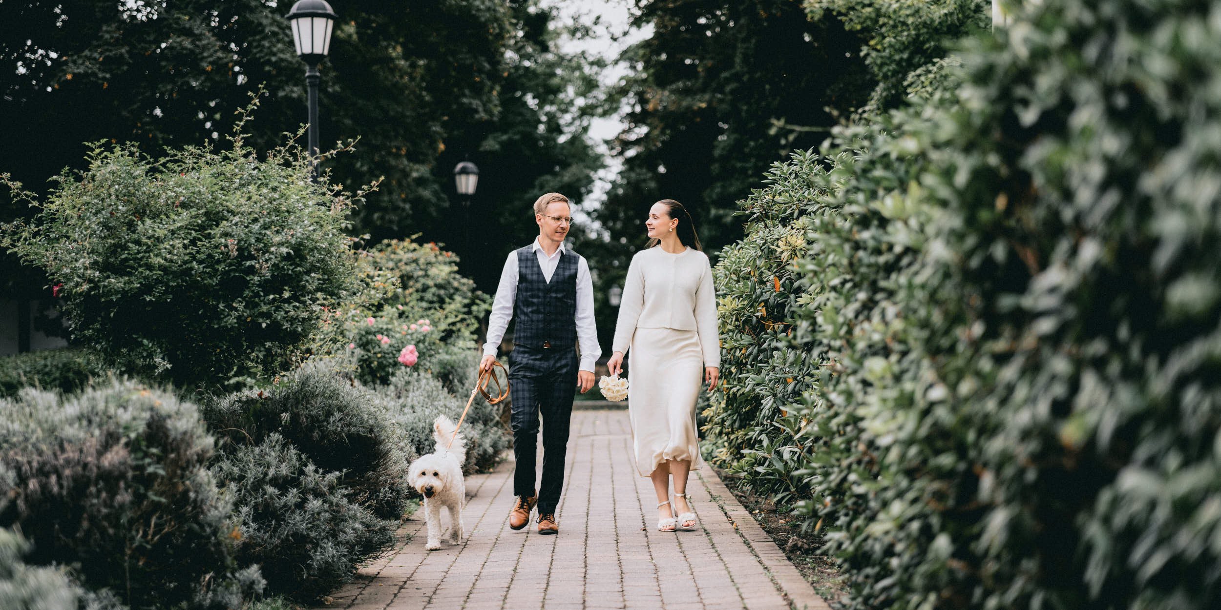 Ein Hochzeitspaar, das zur Trauung im Standesamt im Kloster Wöltingerode spaziert, begleitet von einem weißen Hund und umgeben von grünen Büschen und Bäumen.