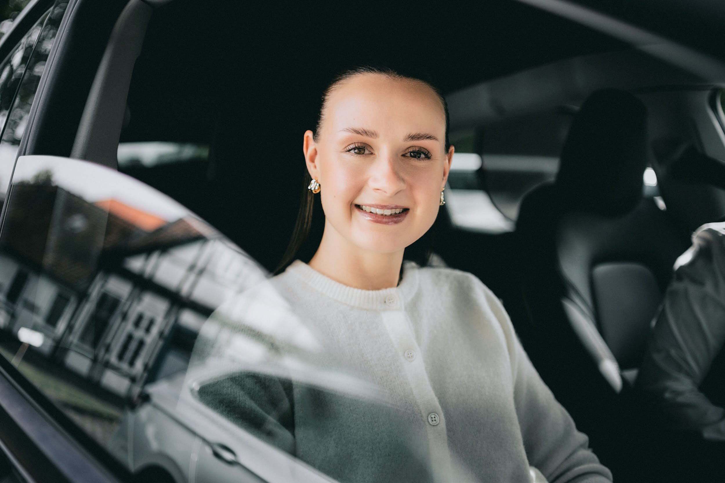 Lächelnde Frau sitzt im Auto, Blick durch die Windschutzscheibe nach vorne, trägt Ohrringe und Pullover.