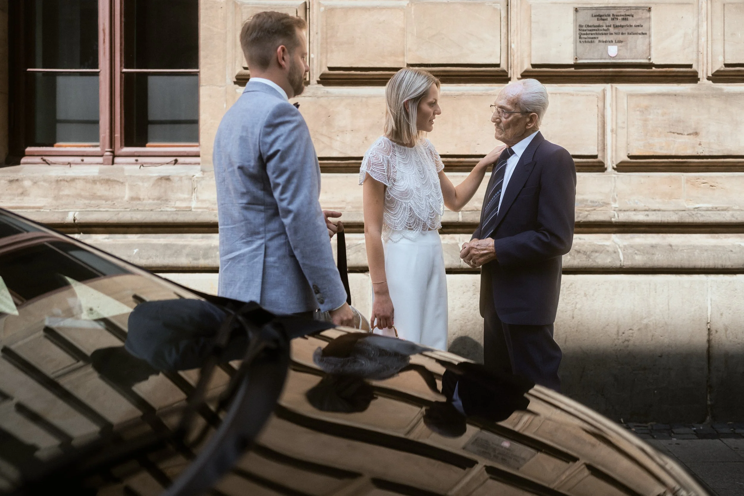 Drei Menschen stehen vor einem historischen Gebäude und führen ein Gespräch. Es handelt sich um eine ältere Frau, einen jüngeren Mann und einen weiteren Mann, die alle formell gekleidet sind.