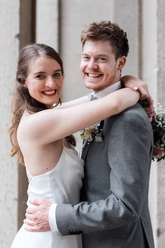 Ein lächelndes Brautpaar bei einer Hochzeit, das sich umarmt, vor einem steinernen Hintergrund.