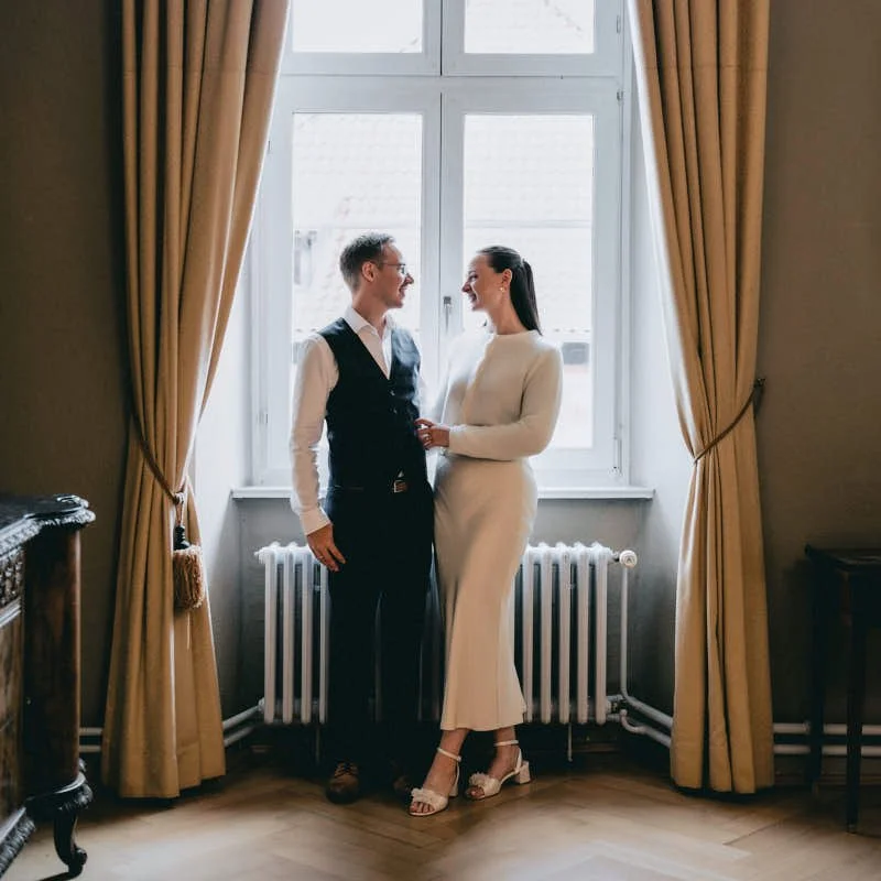 Ein Mann und eine Frau stehen sich vor einer großen Fensterfront gegenüber, lachen und halten sich an den Händen. Sie befinden sich in einem hellen Raum mit beige Vorhängen.