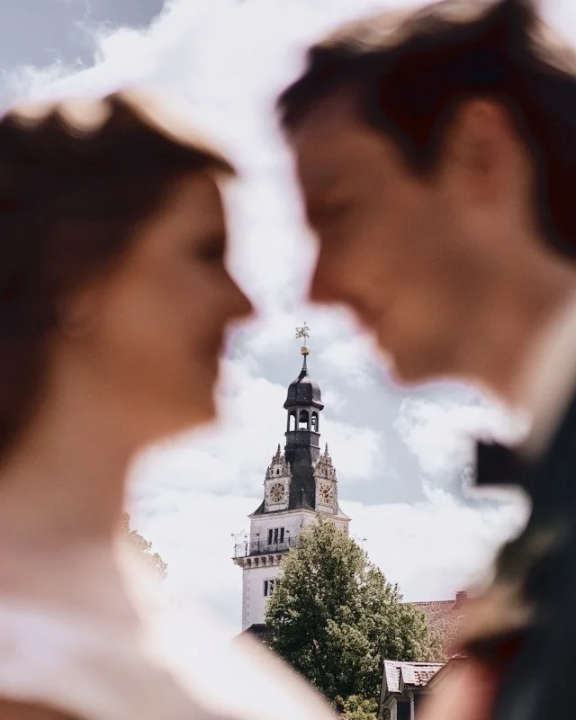 Paare, die sich vor einem historischen Turm küssen, mit einem blauen Himmel und Wolken im Hintergrund.