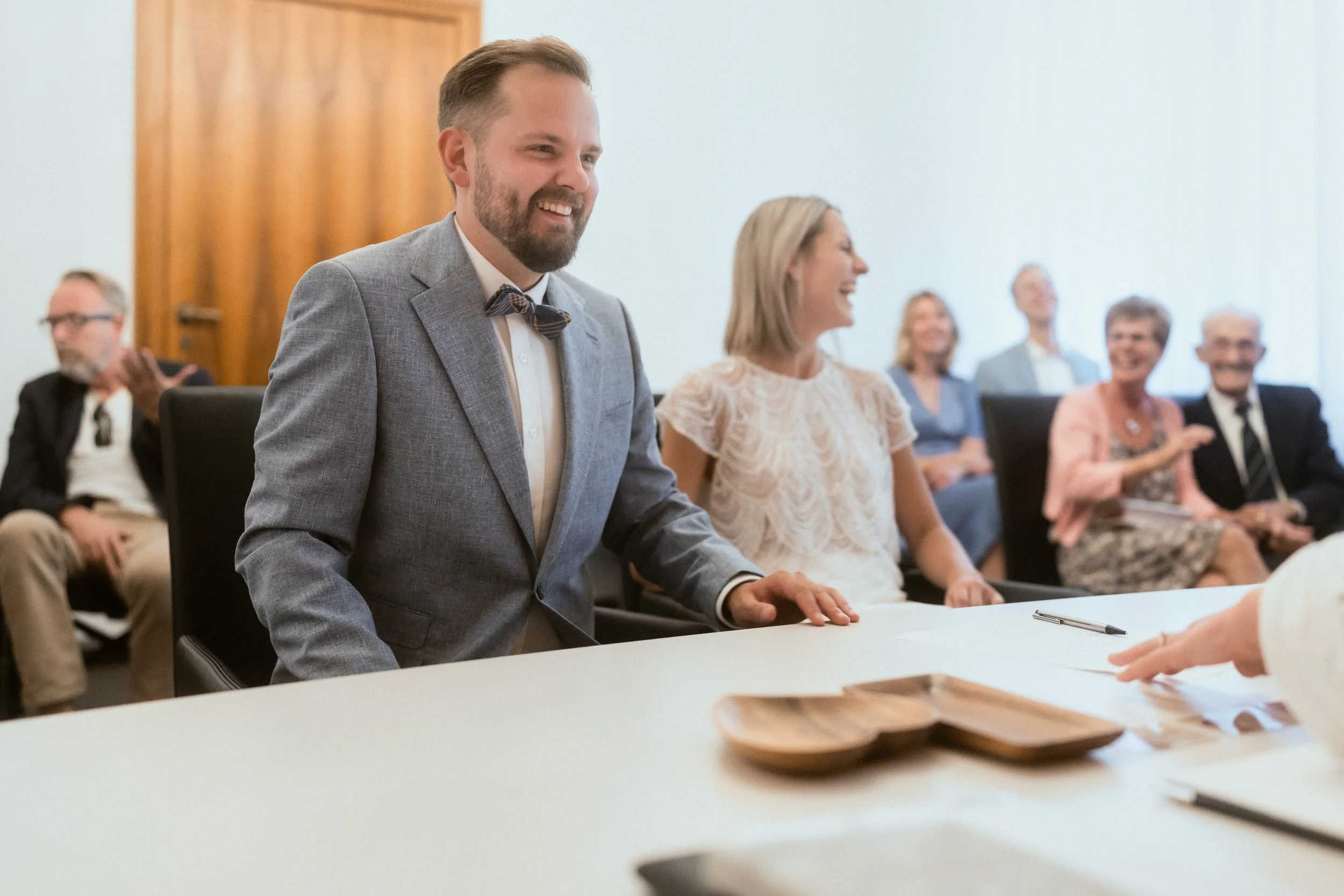 Ein Mann in einem grauen Anzug und Krawatte sitzt an einem Tisch, umgeben von lachenden Menschen bei einer Feier oder einem Treffen in einem Raum mit weißem Wand und Holzdekor.