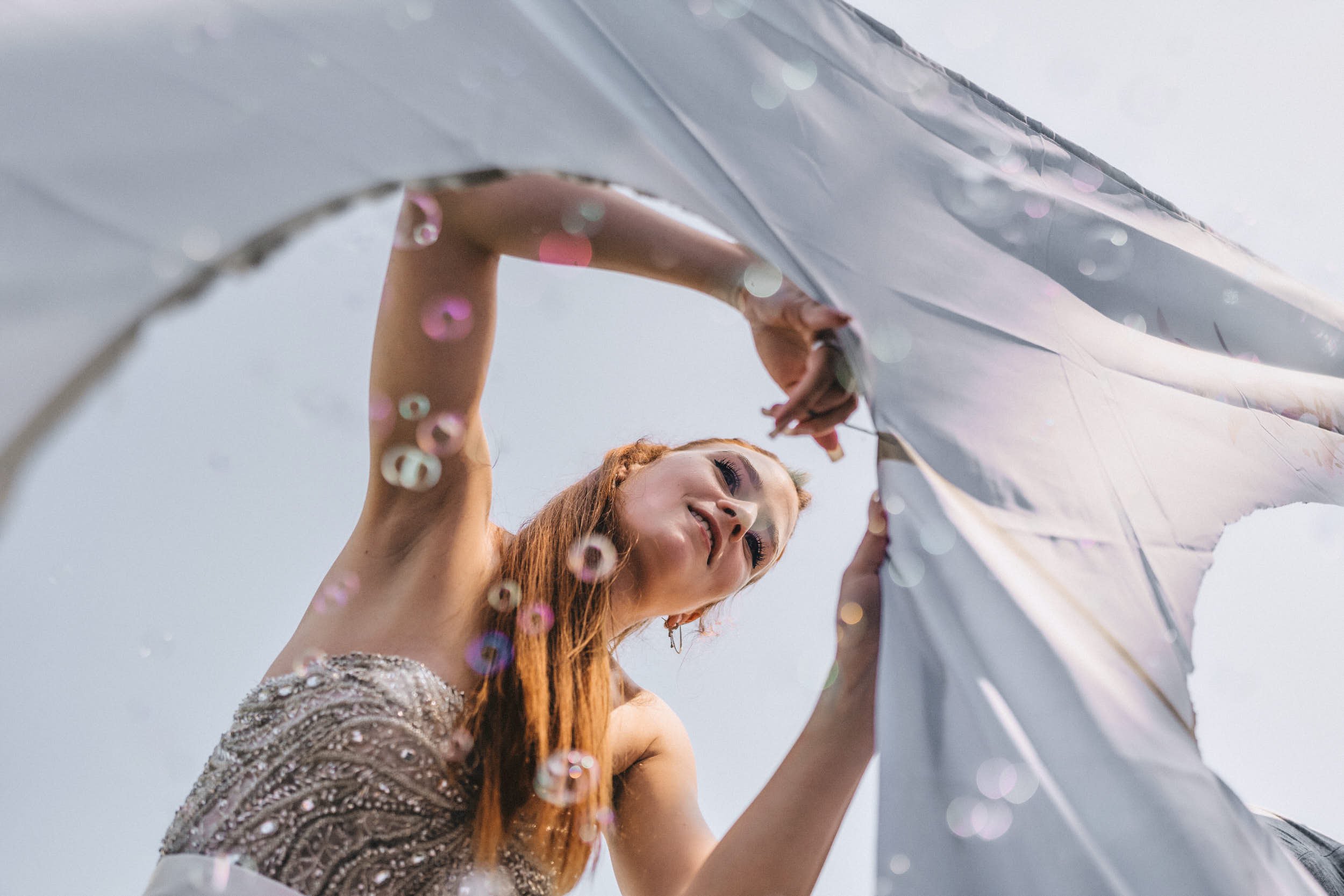 Frau mit rotem Haar und einem glänzenden, silbernen Kleid lächelt bei einem Fotoshooting mit einem Reflektor im Freien, Sonnenlicht im Hintergrund.