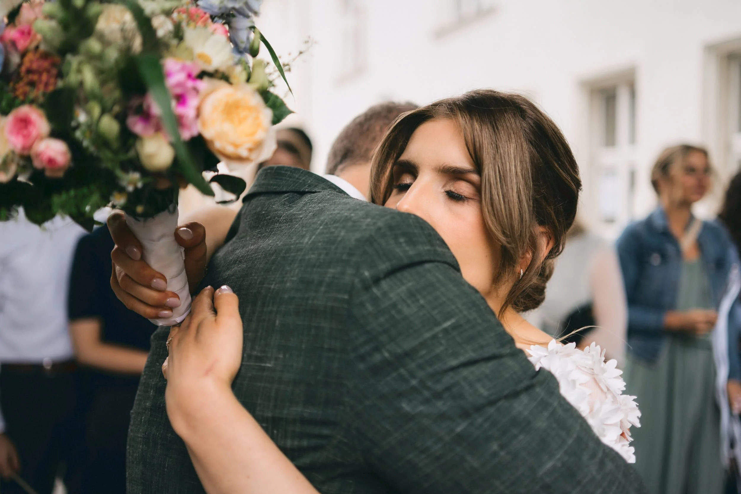 Ein Mann und eine Frau umarmen sich emotional bei einer Feier, die Frau hält einen Blumenstrauß.