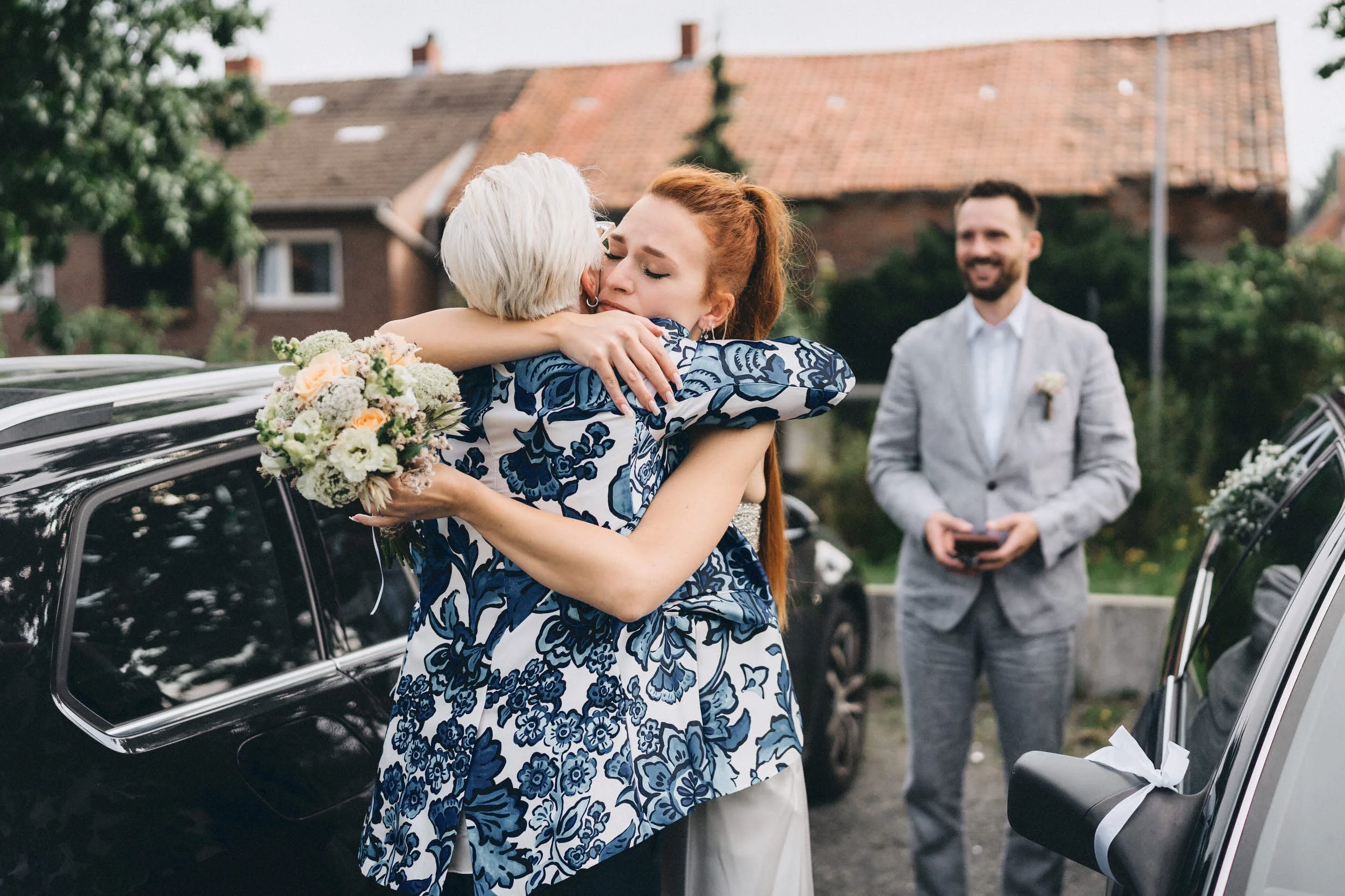Eine Frau umarmt eine ältere Frau mit Blumenstrauß bei einem Familienfest im Freien, während ein Mann im Hintergrund lächelt.