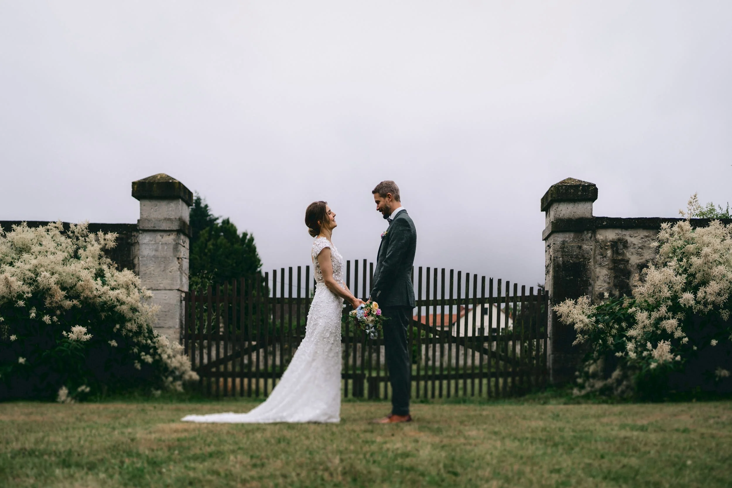 Ein Brautpaar steht sich bei ihrer Hochzeit im Freien in der Mitte eines Gartens gegenüber, hält sich an den Händen und schaut sich verliebt an, während die Braut eine weiße Hochzeitskleidung trägt und der Bräutigam einen dunklen Anzug. Im Hintergrun