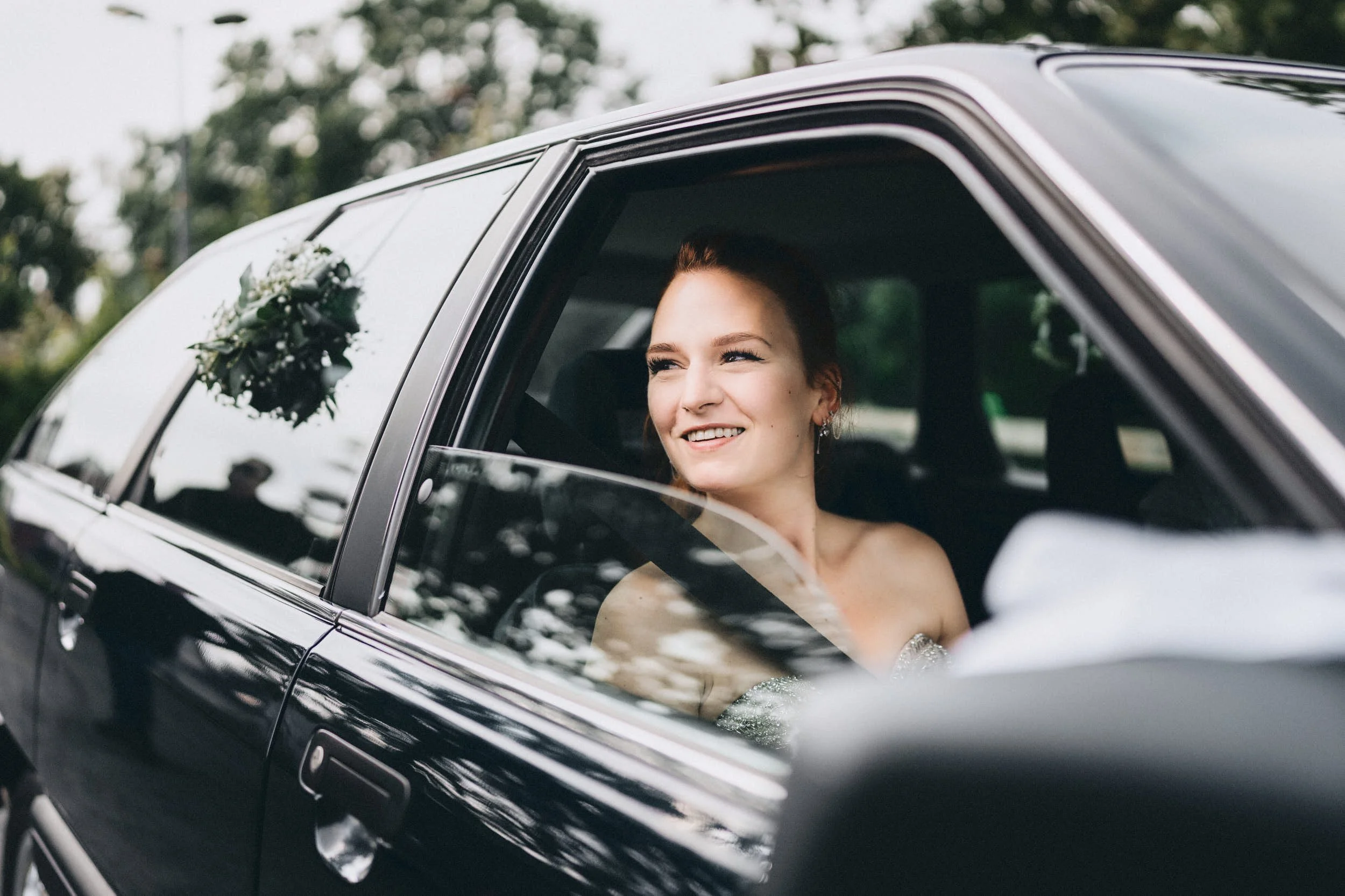 Eine Frau sitzt in einem schwarzen Auto, lächelt und schaut zum Fenster hinaus, während im Hintergrund Bäume zu sehen sind.