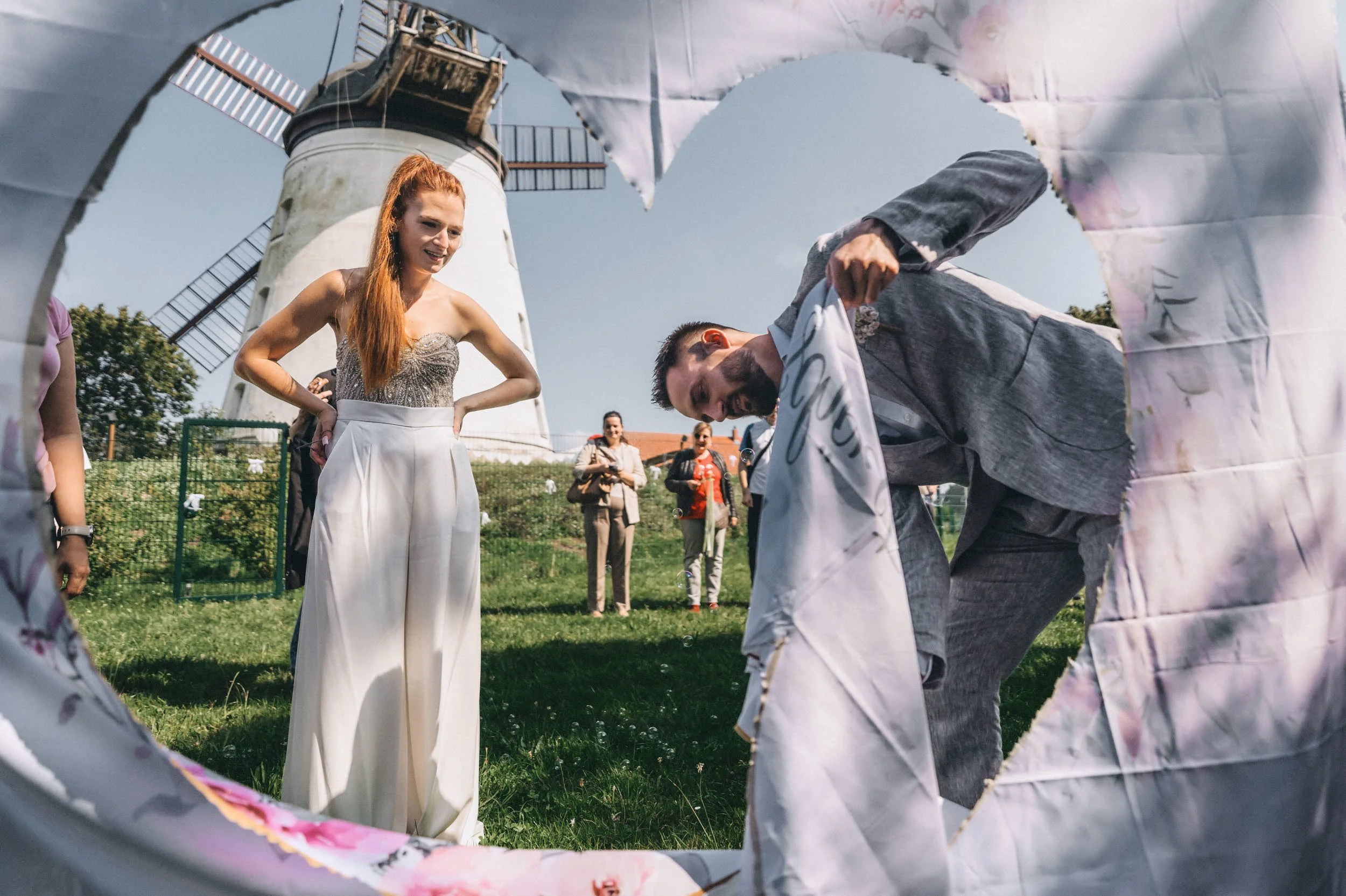 Eine Gruppe von Menschen, darunter eine Frau mit langem roten Haar und einem weißen Kleid, steht im Freien vor einem Leuchtturm, während sie durch ein zerbrochenes Herz aus Papier oder Pappe schauen, das im Vordergrund liegt.