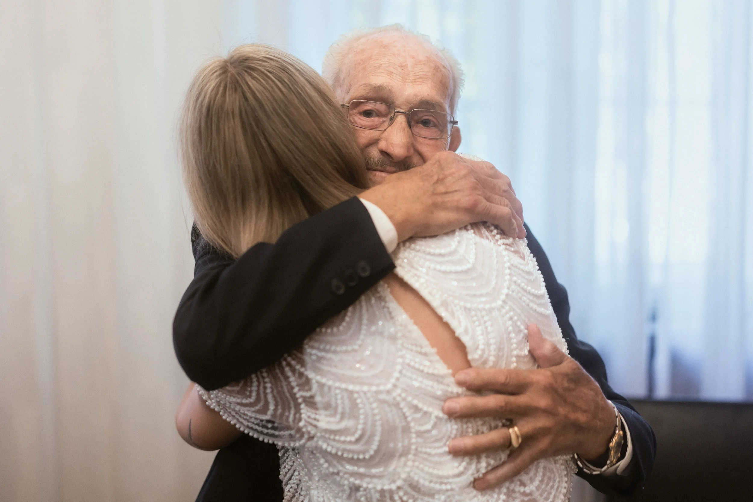 Ein älterer Mann und eine Frau umarmen sich herzlich. Der Mann trägt eine Brille und einen dunklen Anzug, die Frau trägt ein weißes Kleid mit Perlen und hat lange, blonde Haare.