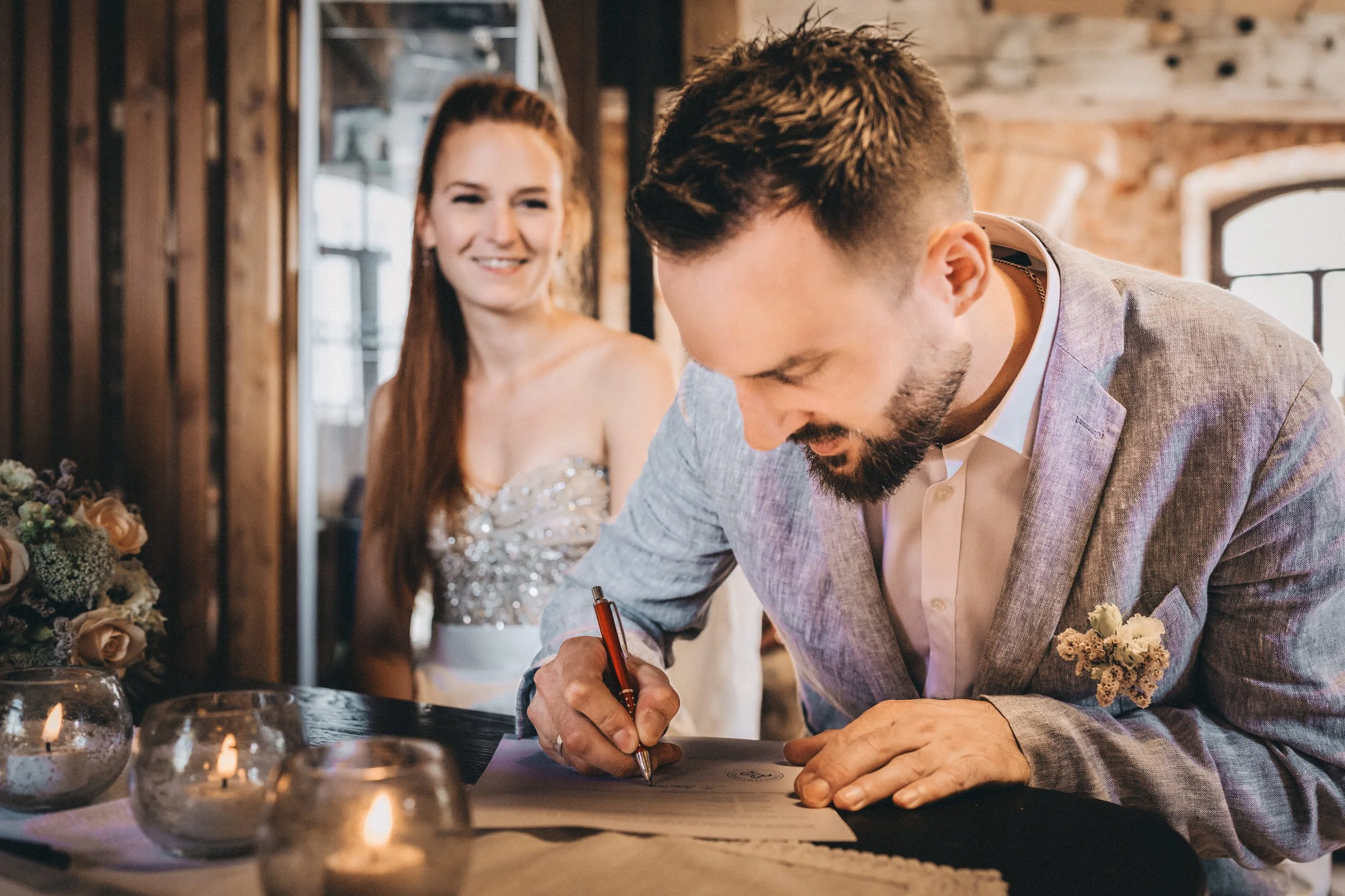 Ein Mann unterschreibt ein Dokument bei einer Hochzeit, während eine Braut im Hintergrund lächelt. Es gibt Kerzen auf dem Tisch.