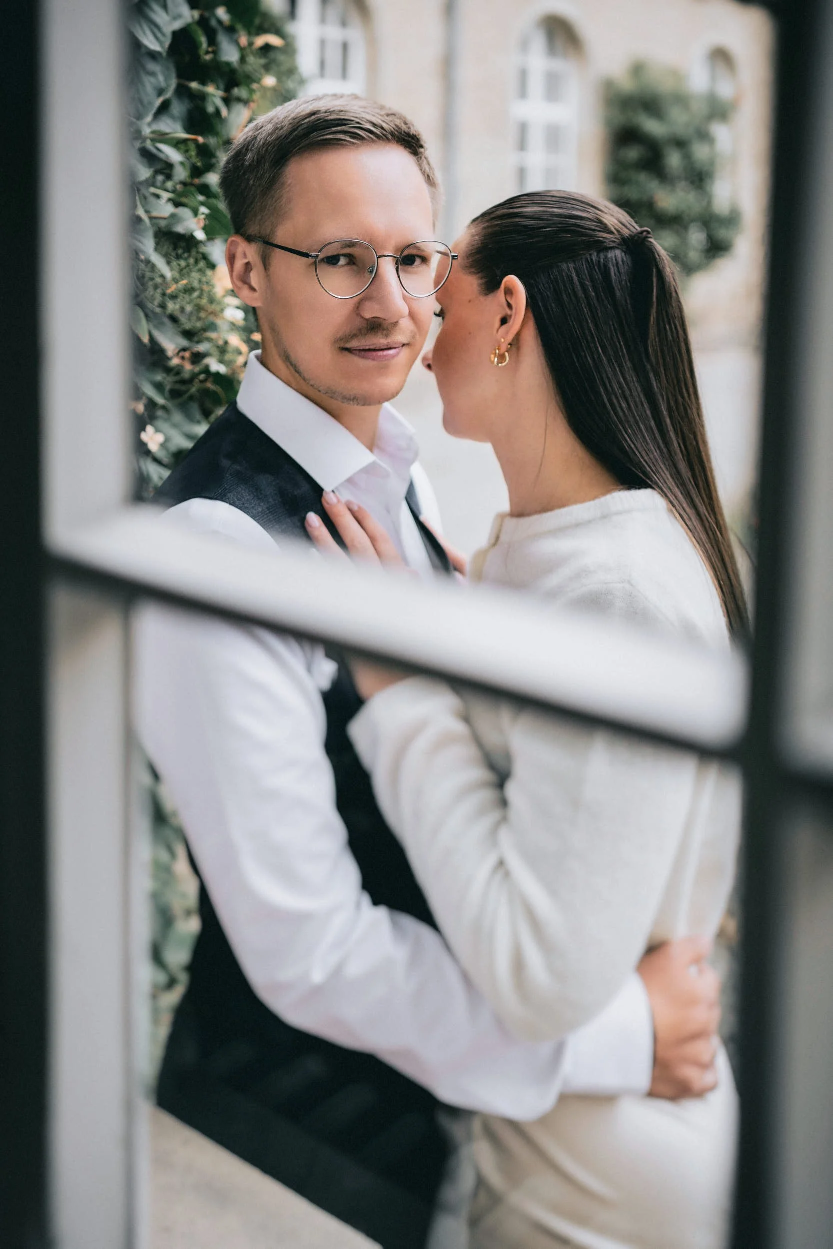 Ein Mann trägt eine Brille, ein weißes Hemd und eine dunkle Weste, eine Frau trägt eine cremefarbene Jacke und hat dunkle, lange Haare, sie stehen sich sehr nah, ihre Gesichter sind fast aneinander, während sie sich im Freien vor einem Gebäude befind