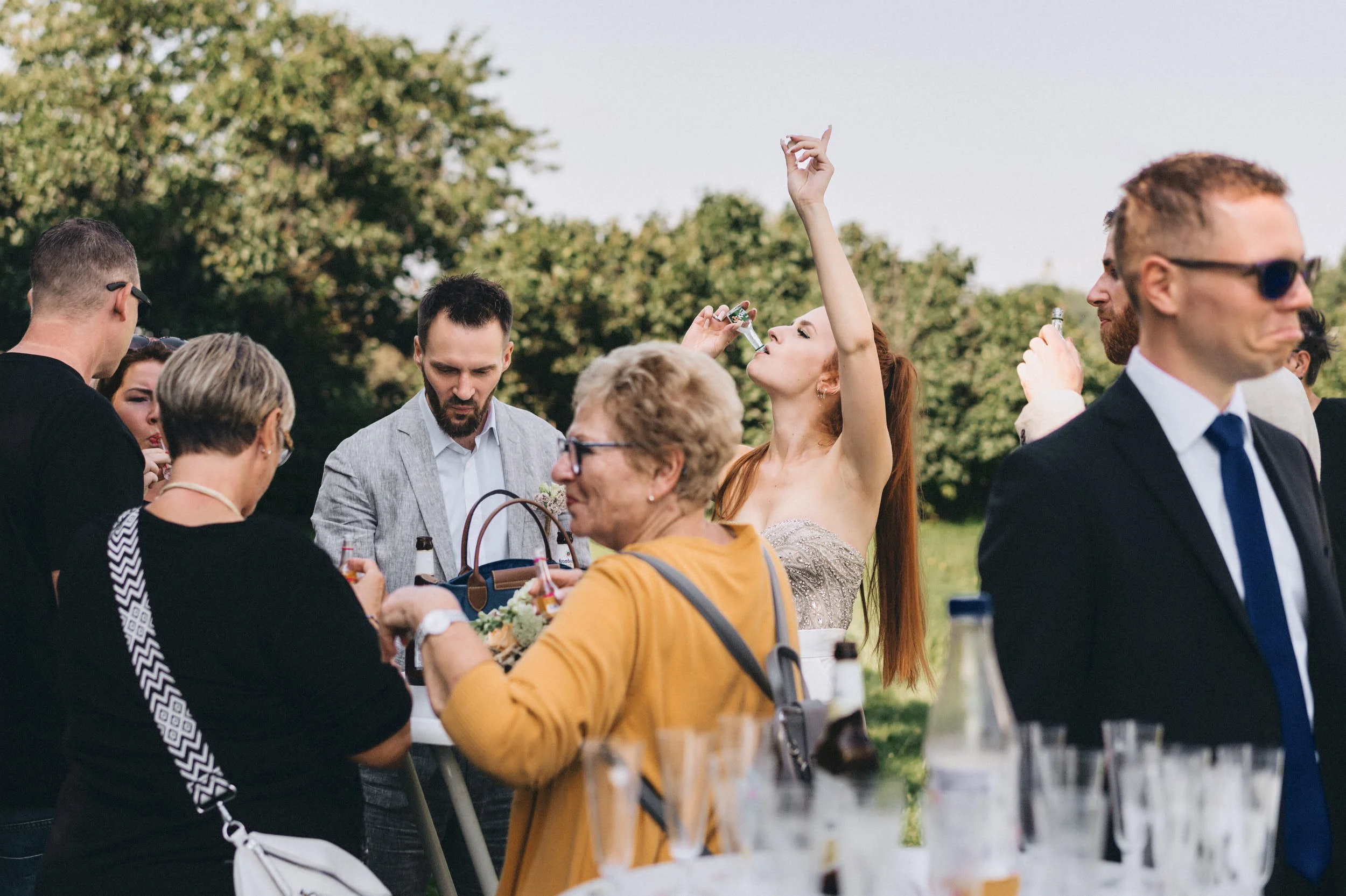 Hochzeit im Freien mit mehreren Gästen, einige trinken, eine Frau hebt die Hand hoch, andere unterhalten sich, im Hintergrund Bäume und ein blauer Himmel.