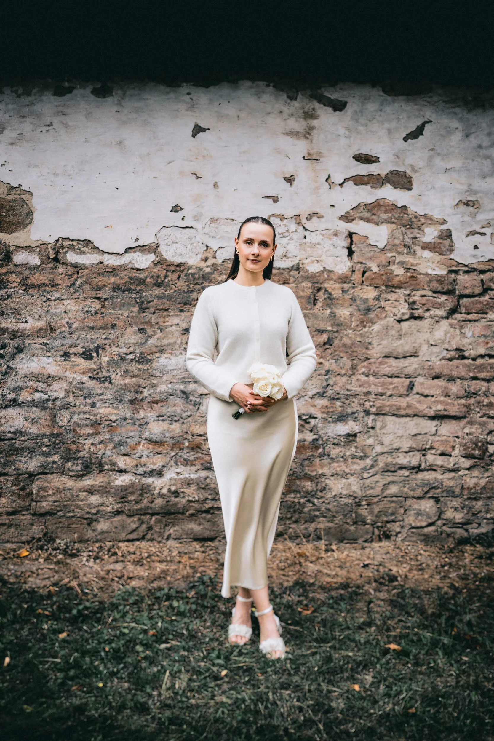 Eine Frau in einem eleganten weißen Kleid mit Blumen in den Händen steht vor einer alten, abgeblätterten Steinmauer.