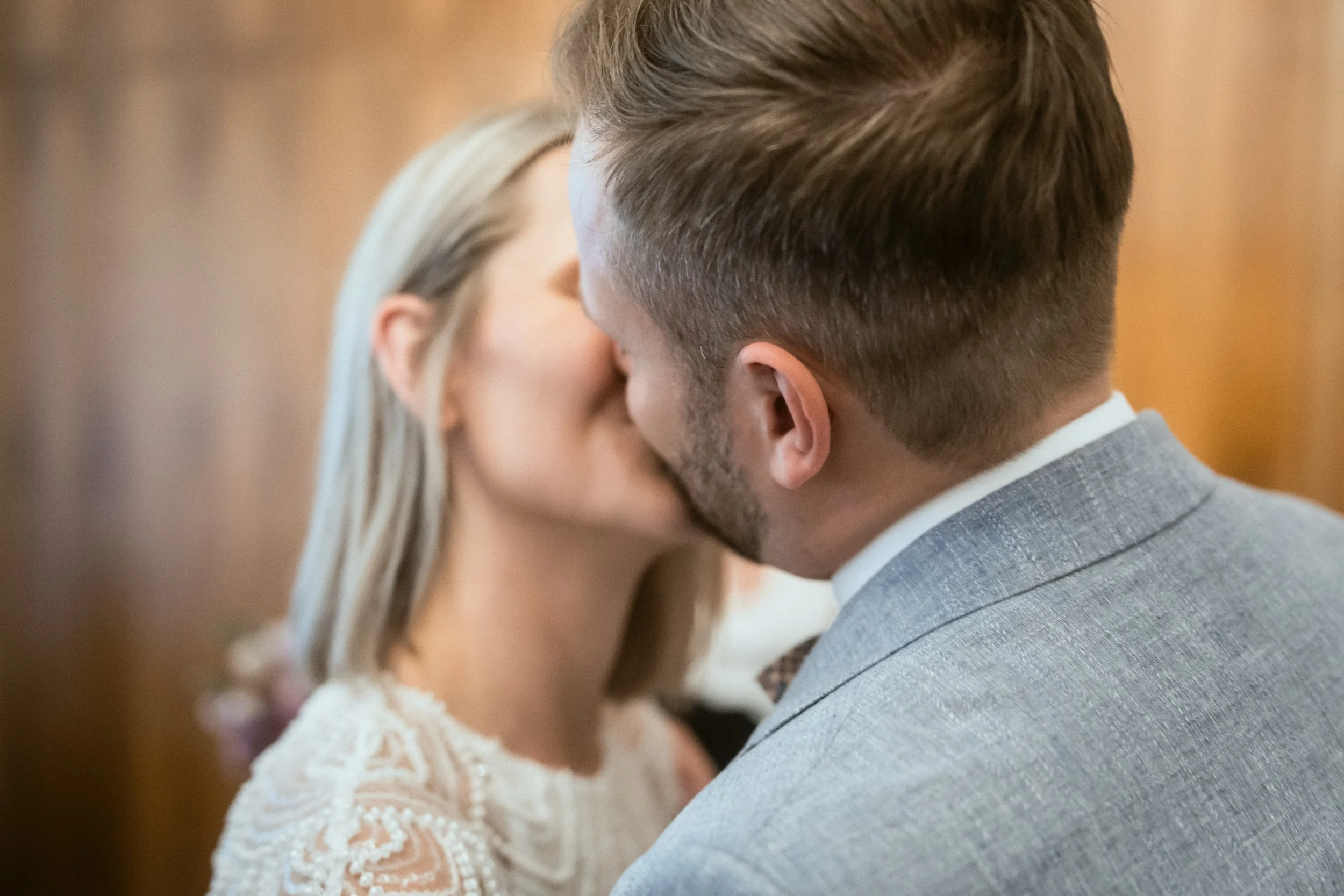 Ein Mann und eine Frau küssen sich in einer Nähe, die beide fast ihren ganzen Gesichter zeigt. Der Mann trägt einen grau karierten Anzug, die Frau eine weiße Spitze-Kleid. Der Hintergrund ist unscharf.