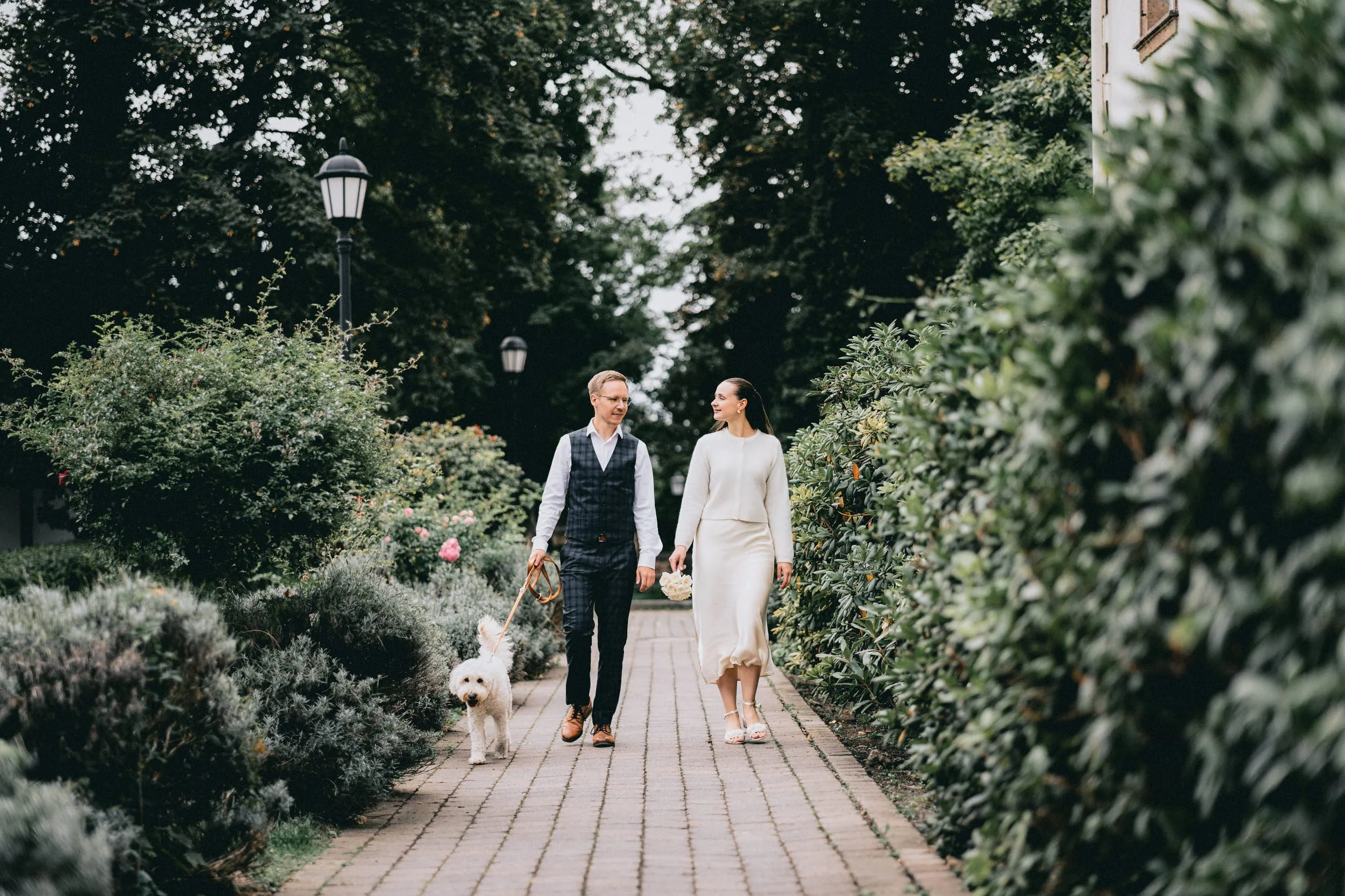 Ein frisch verheiratetes Paar geht Hand in Hand durch einen gepflegten Garten, begleitet von einem weißen Hund, während es sich mit einer Blumenstrauß in der Hand zueinander bewegt.