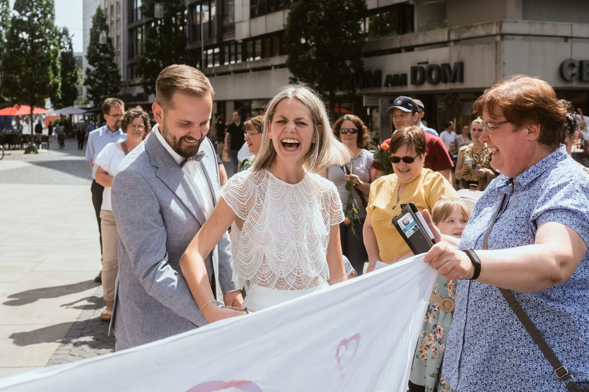 Menschen bei einer Outdoor-Veranstaltung, wahrscheinlich eine Hochzeit, mit einem Brautpaar, das von mehreren Personen unterstützt wird, während sie eine Banner oder ein Banner hält.