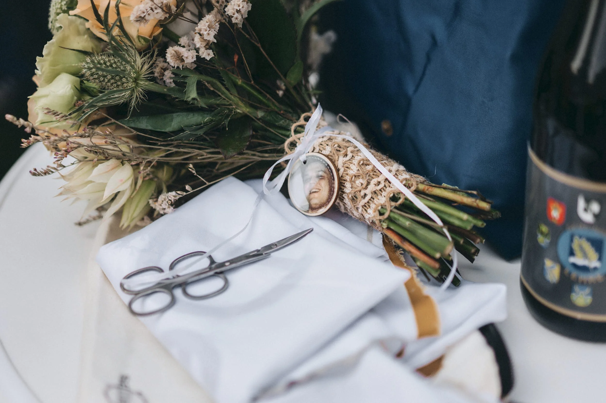 Ein Blumenstrauß mit weißen Rosen, getrockneten Blumen und Gräsern liegt auf einem weißen Tuch. An dem Strauß hängt eine Medaille mit einem lachenden Gesicht.