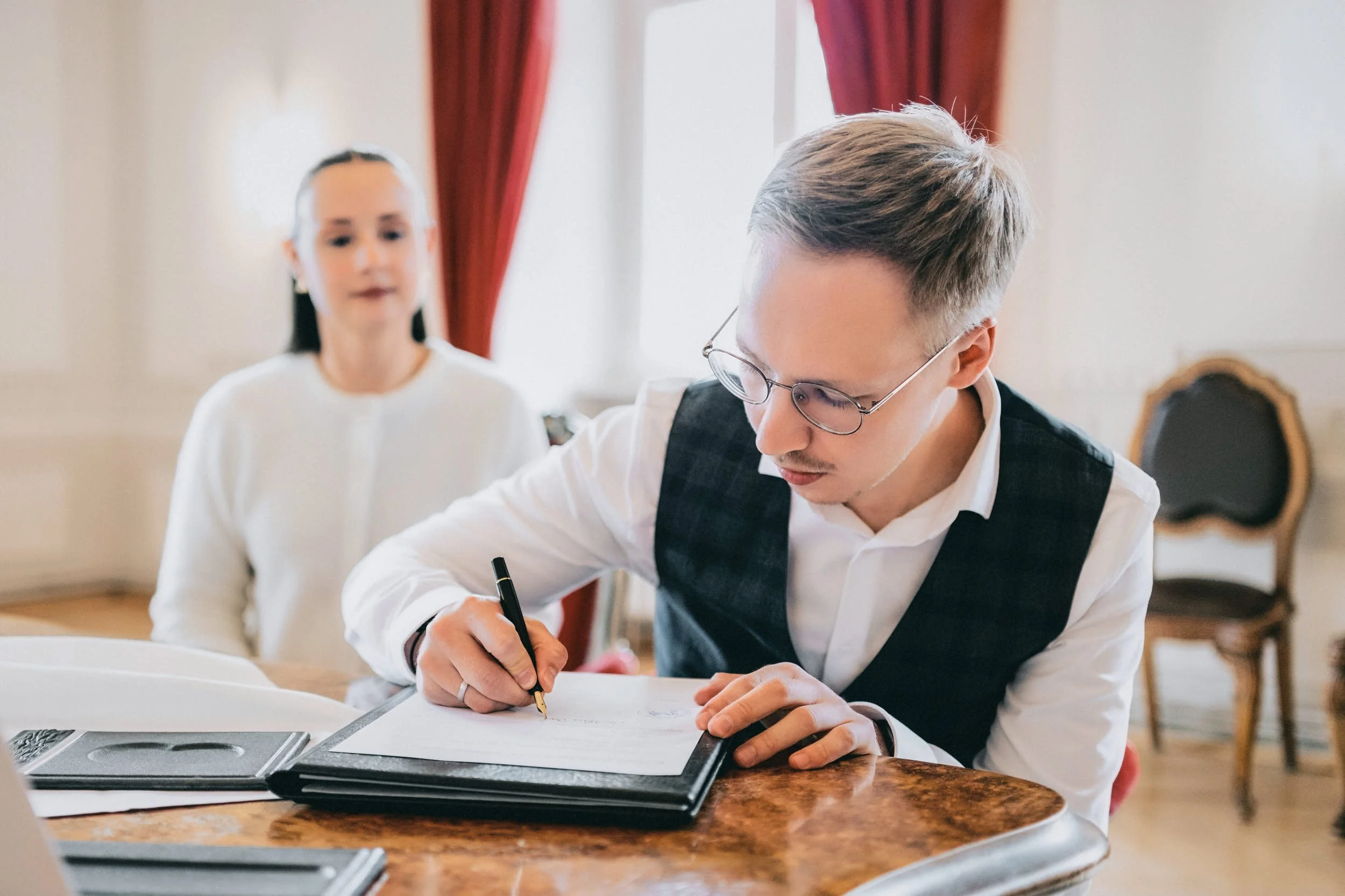 Ein junger Mann mit Brille schreibt auf einem Dokument auf einem Tisch. Im Hintergrund eine junge Frau, die zuschaut. Der Raum ist elegant mit roten Vorhängen und großen Fenstern.