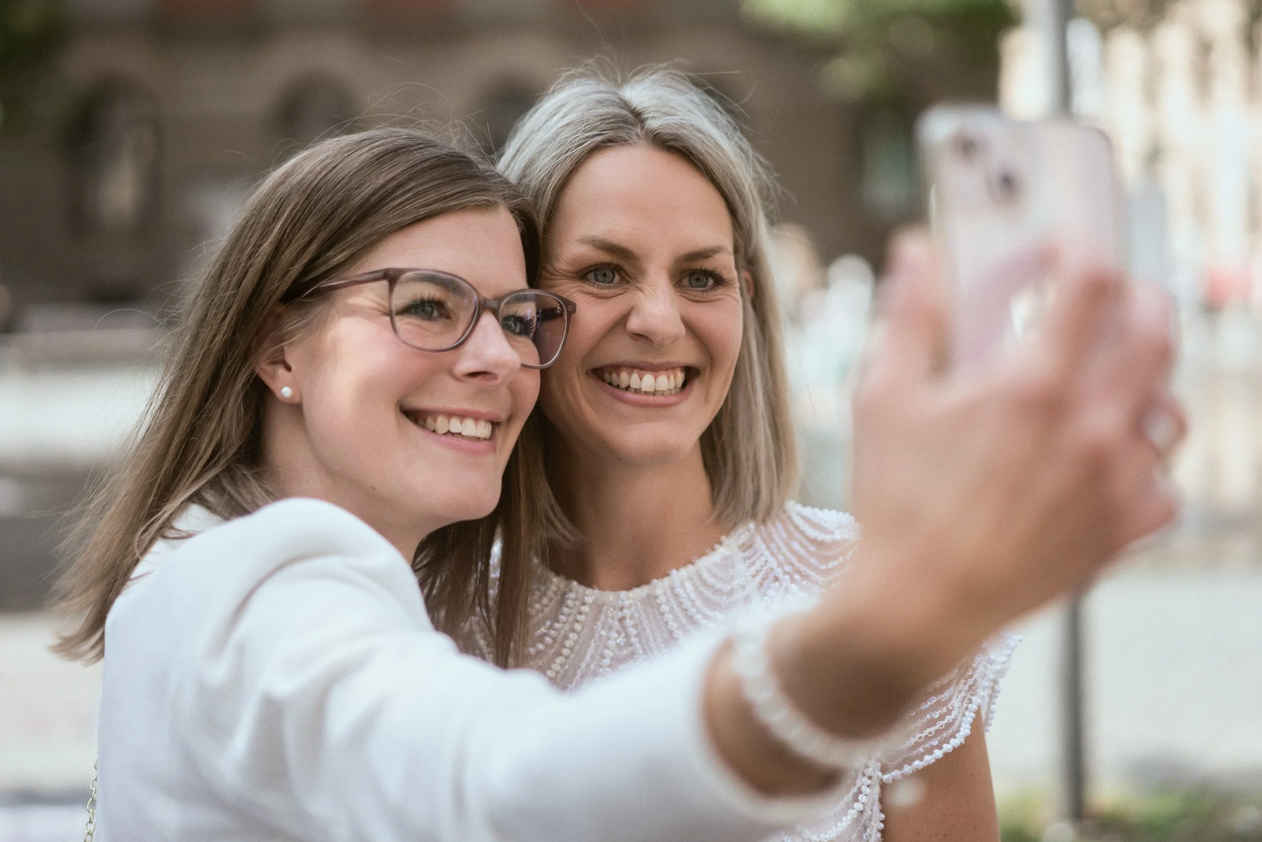 Zwei Frauen machen ein Selfie im Freien, beide lächeln und haben graues oder braunes Haar, eine trägt Brille, die andere hat lockiges blondes Haar.