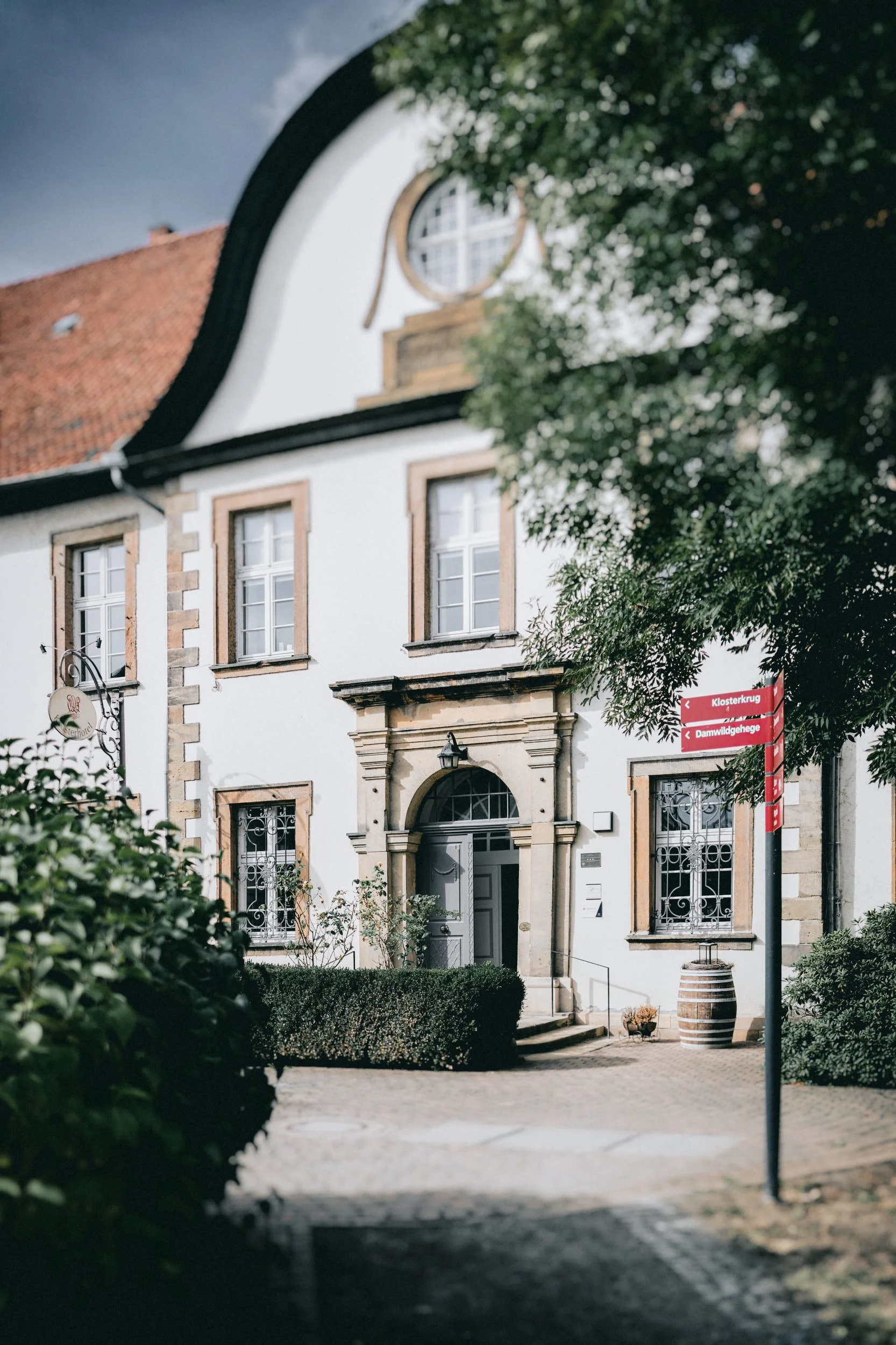Ein georgianisches Gebäude mit weißen Wänden, braunen Fensterrahmen und einer klassischen Tür. Es gibt einen Eingang mit Treppe, einen Zaun, einen Baum und eine Straßenlaterne daneben. Ein rotes Straßenschild zeigt den Weg zu einem Klosterkrug und Da