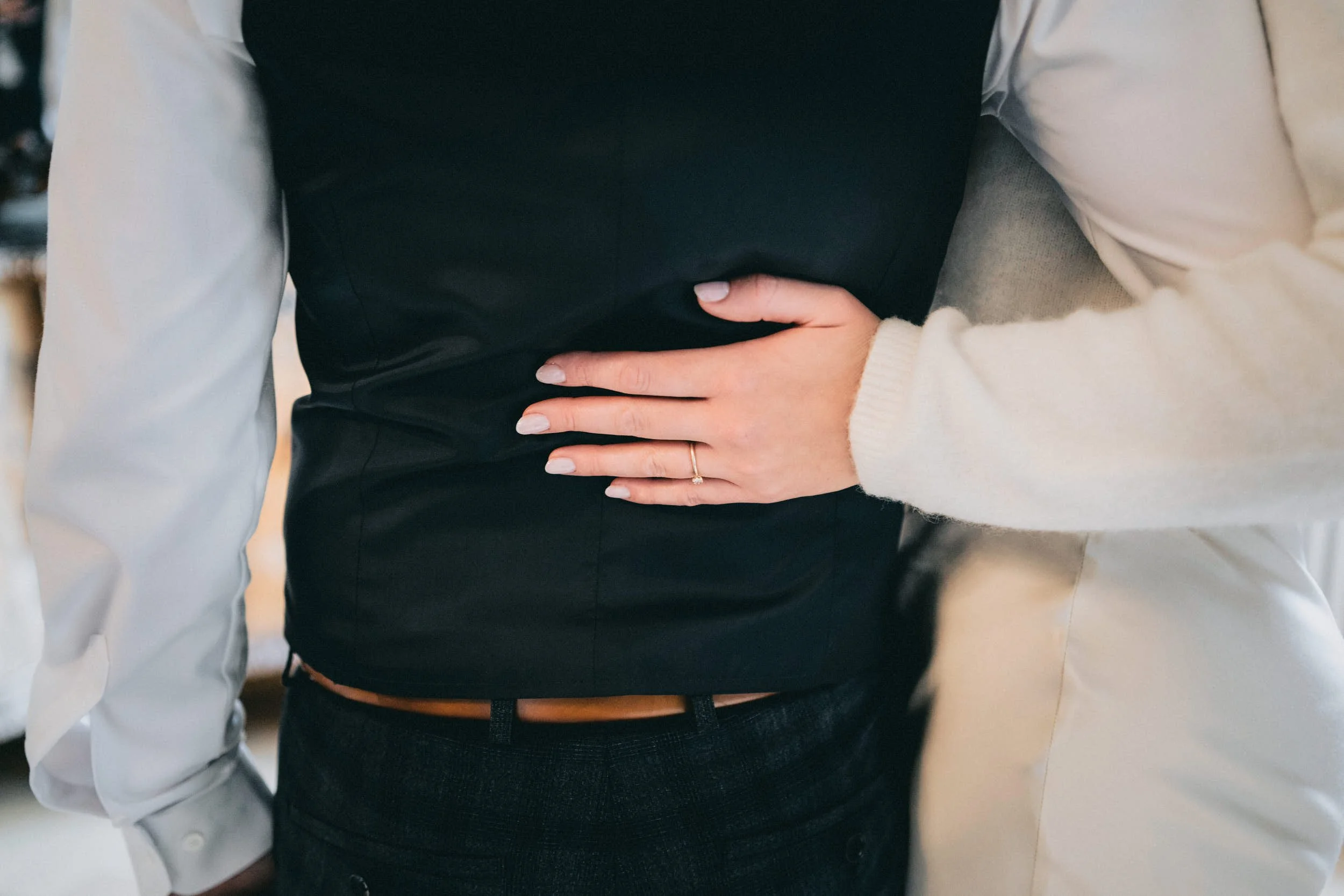 Nahaufnahme von zwei Personen, eine hält ihre Hand auf die Taille der anderen, beide tragen elegante Kleidung. Die Hand der Person auf der rechten Seite trägt einen goldenen Ring.