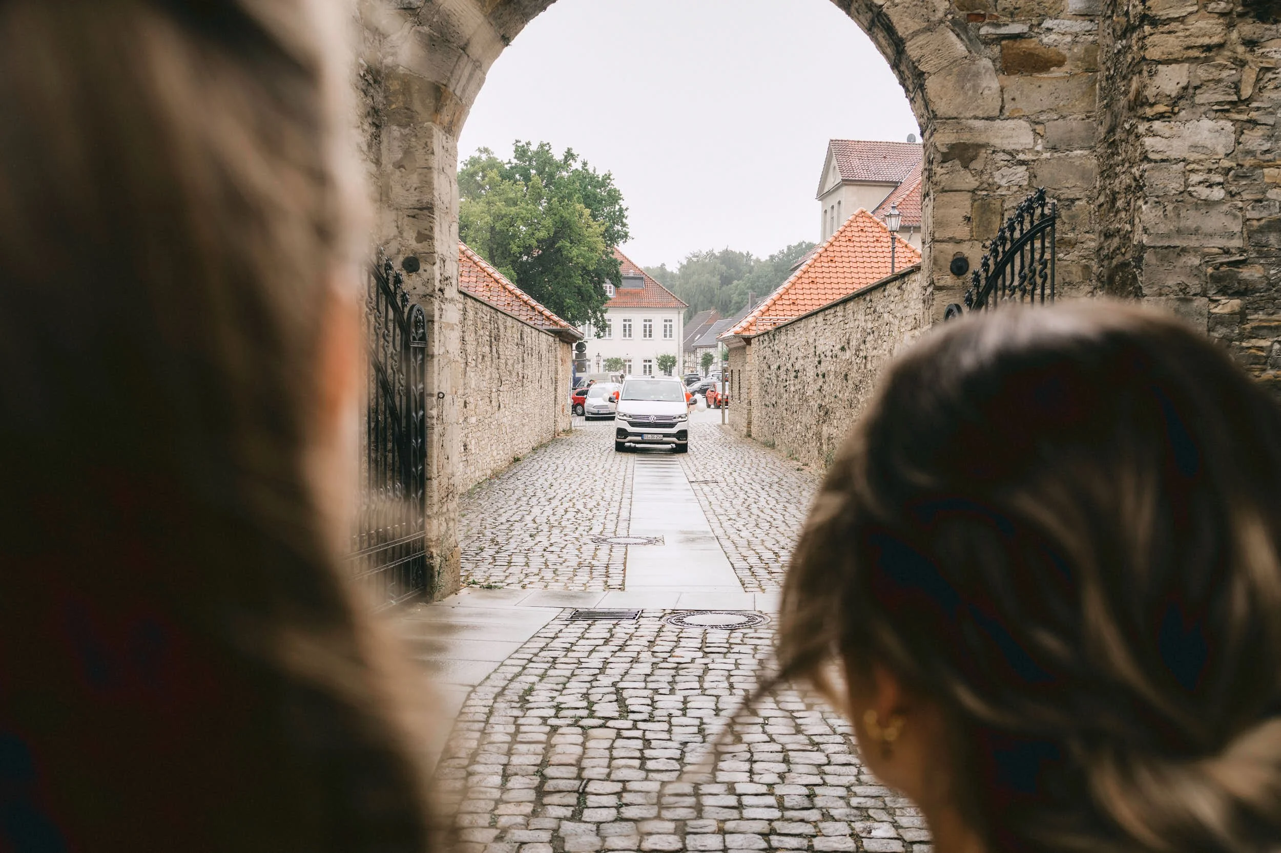 Blick durch einen Torbogen auf eine kopfsteinpflasterte Straße mit Steinmauer, Parkende Autos und Gebäude im Hintergrund. Zwei Frauen schauen auf die Szene, ihre Köpfe sind nah am Kameraobjektiv.
