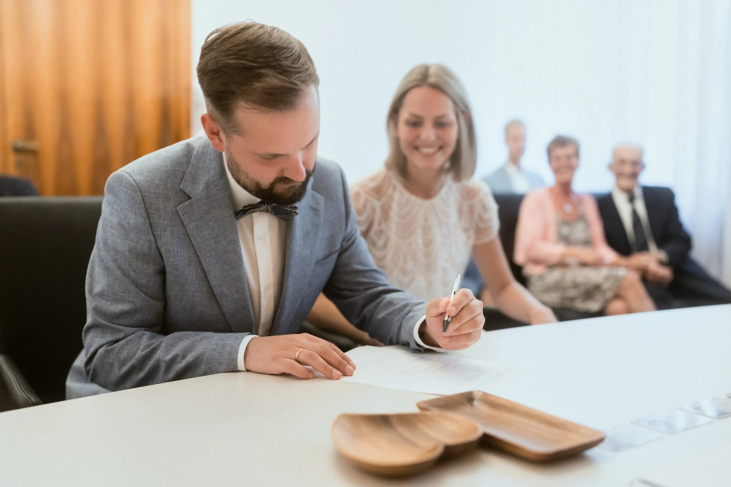 Ein Mann in einem grauen Anzug und mit Fliege schreibt bei einer Sitzung an einem weißen Tisch, im Hintergrund sitzen fünf ältere Menschen, alle in formeller Kleidung, in einem Konferenzraum mit weißen Wänden und Vorhängen.