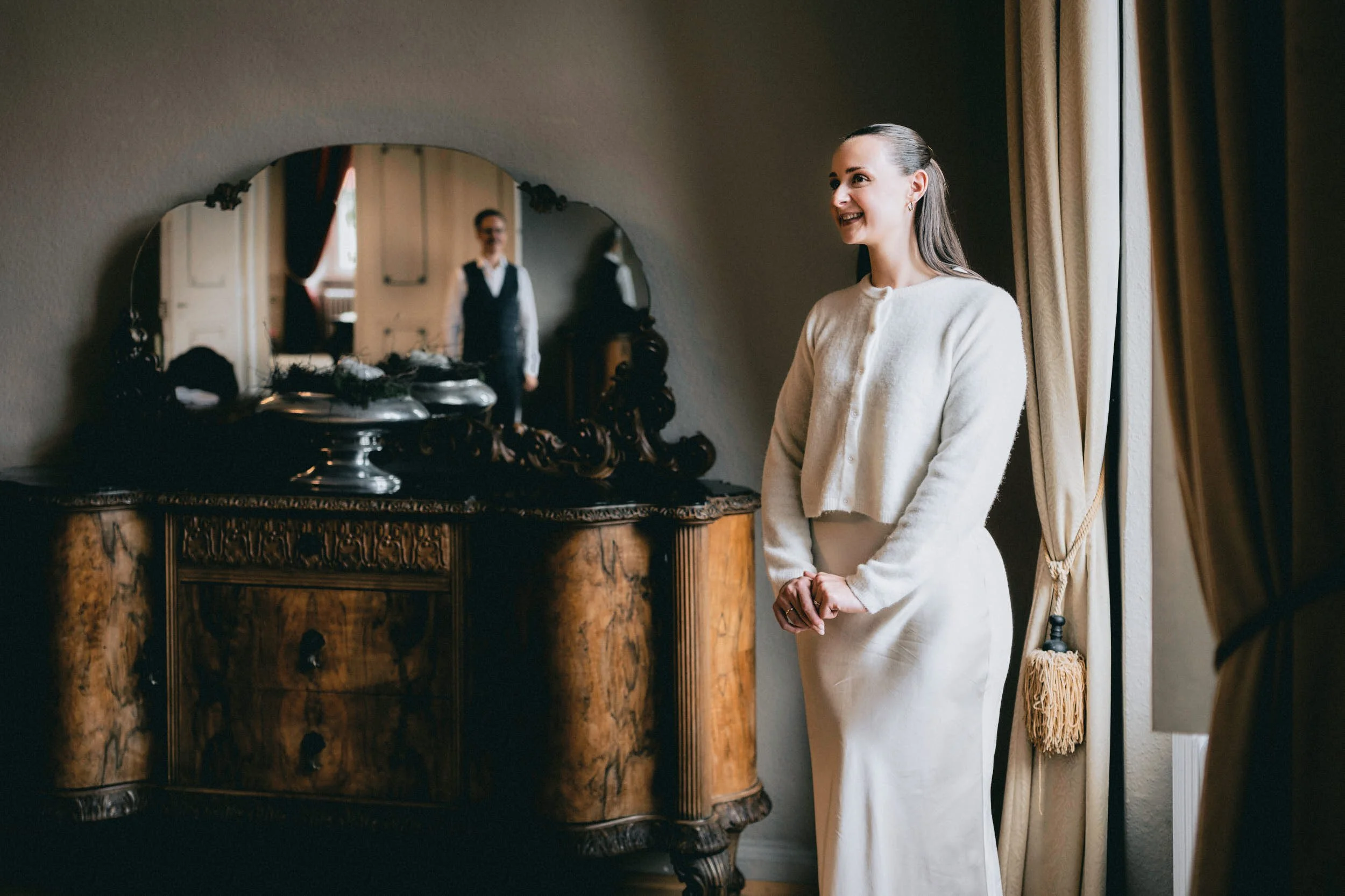 Eine lächelnde Frau steht neben einem antiken Holztisch mit einem Spiegel über dem Tisch, während eine weitere Person im Spiegel zu sehen ist. Die Szene befindet sich in einem elegant eingerichteten Raum.