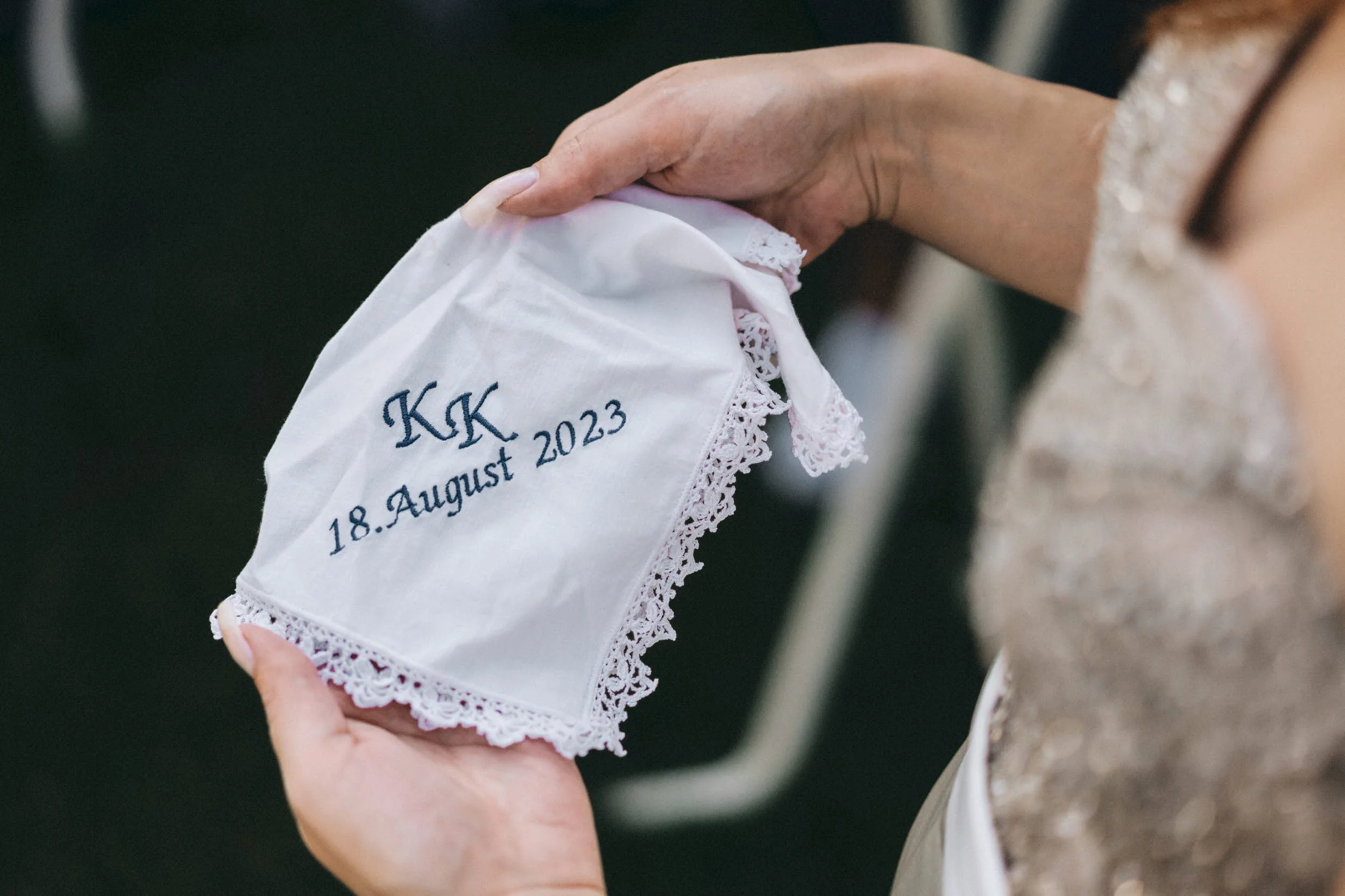 Eine Person hält ein weißes, mit blauer Schrift besticktes Tuch mit Spitzenrändern, auf dem die Initialen 'KK', das Datum '18. August 2023', und weitere Textdetails stehen.