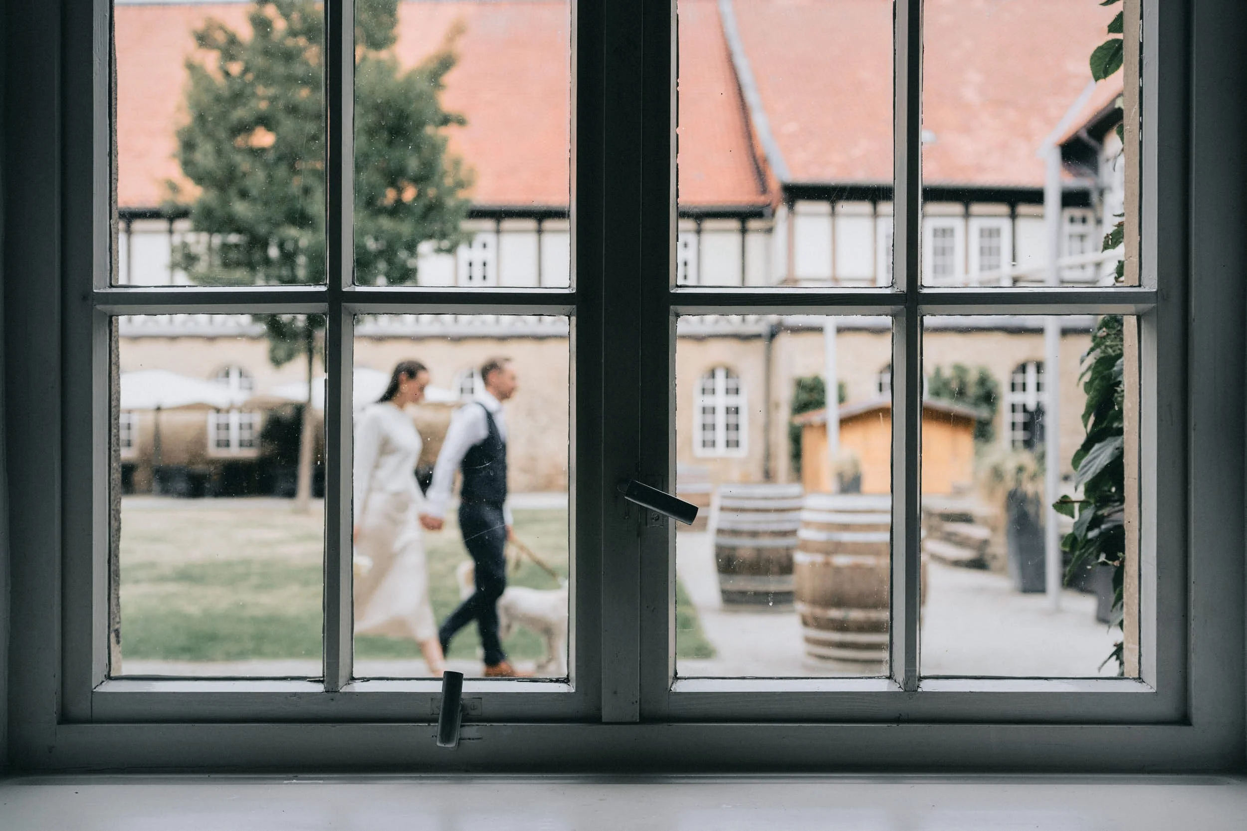 Blick durch ein Fenster auf ein Paar, das auf einer Wiese im Hintergrund spazieren geht, mit Blick auf angrenzende alte Häuser.