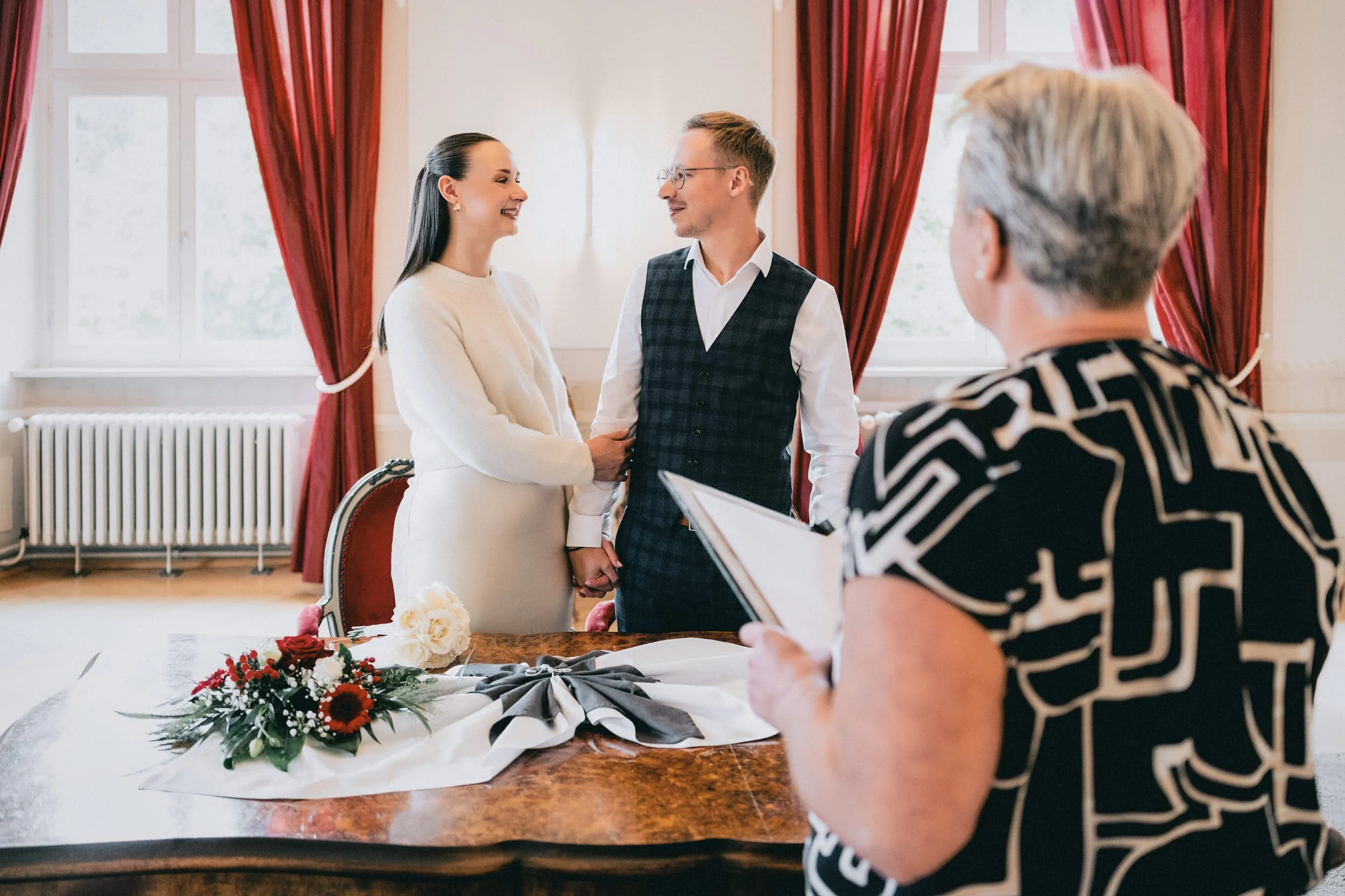 Ein Hochzeitspaar hält sich während der Zeremonie an den Händen, während eine offizielle Person die Heirat bestätigt. Im Vordergrund liegt ein Blumenstrauß auf einem Holztisch.