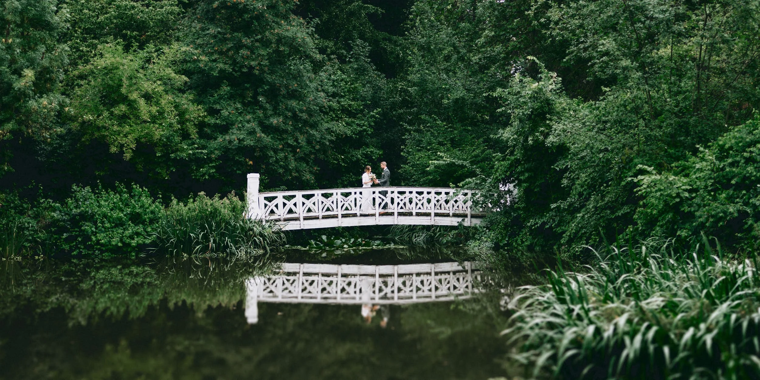 Ein Brautpaar beim Brautpaar-Shooting auf einer weißen Brücke in einem grünen, bewaldeten Park mit Wasser darunter.