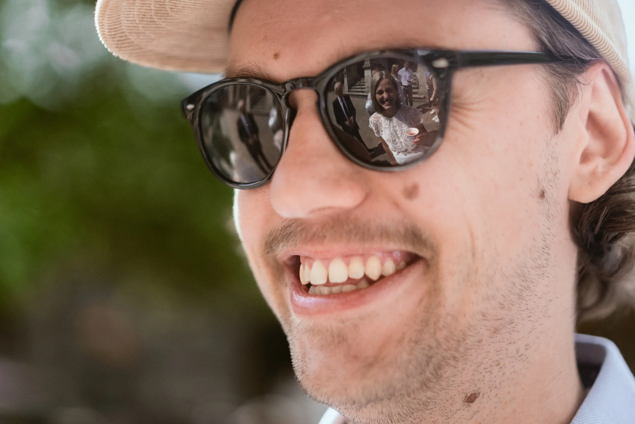 Nahaufnahme eines lachenden Mannes mit Brille und Sonnenhut, der in die Kamera lächelt. Im Sonnenreflex spiegeln sich Menschen, die auf einer Treppe stehen, darunter eine Frau in einem weißen Kleid, die eine Tasse hält.