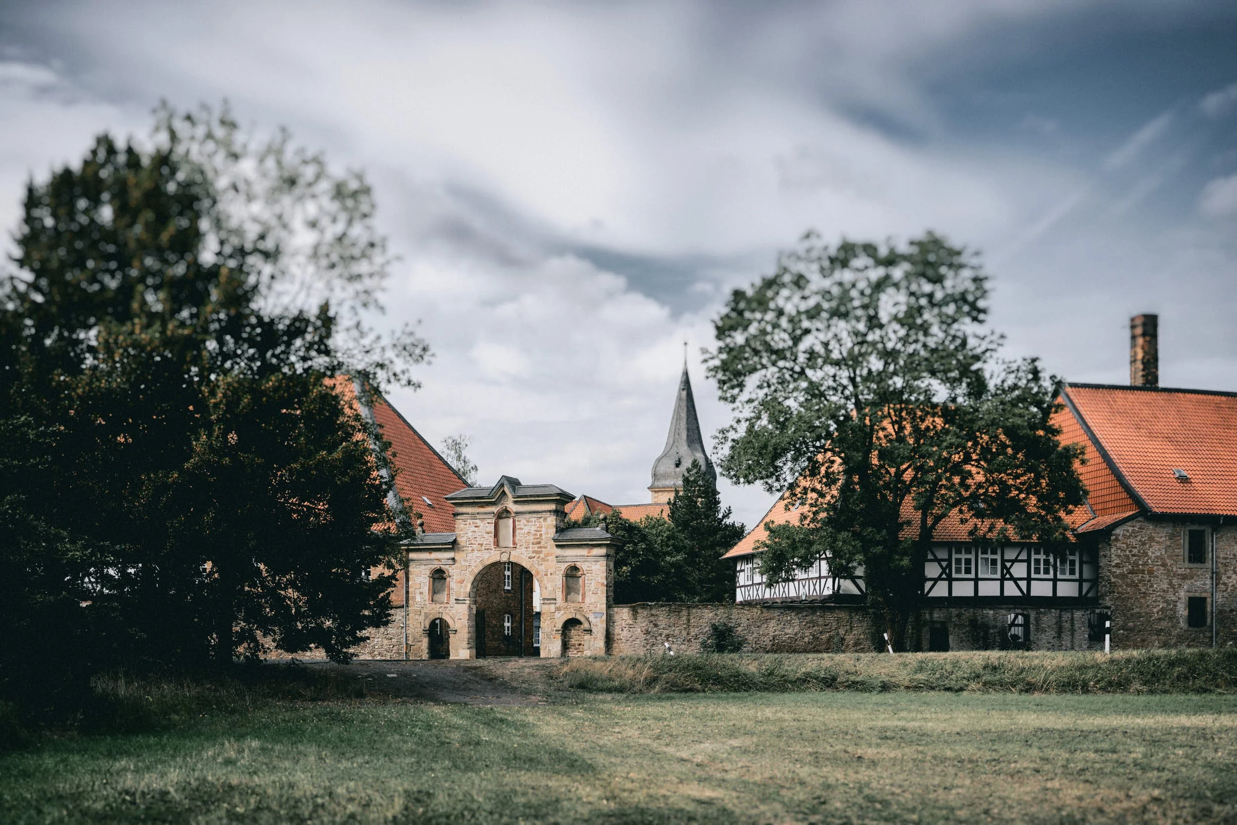 Ein historisches Gebäude mit einem Torbogen, umgeben von grünen Bäumen und roten Dächern, unter einem dunklen, bewölkten Himmel.
