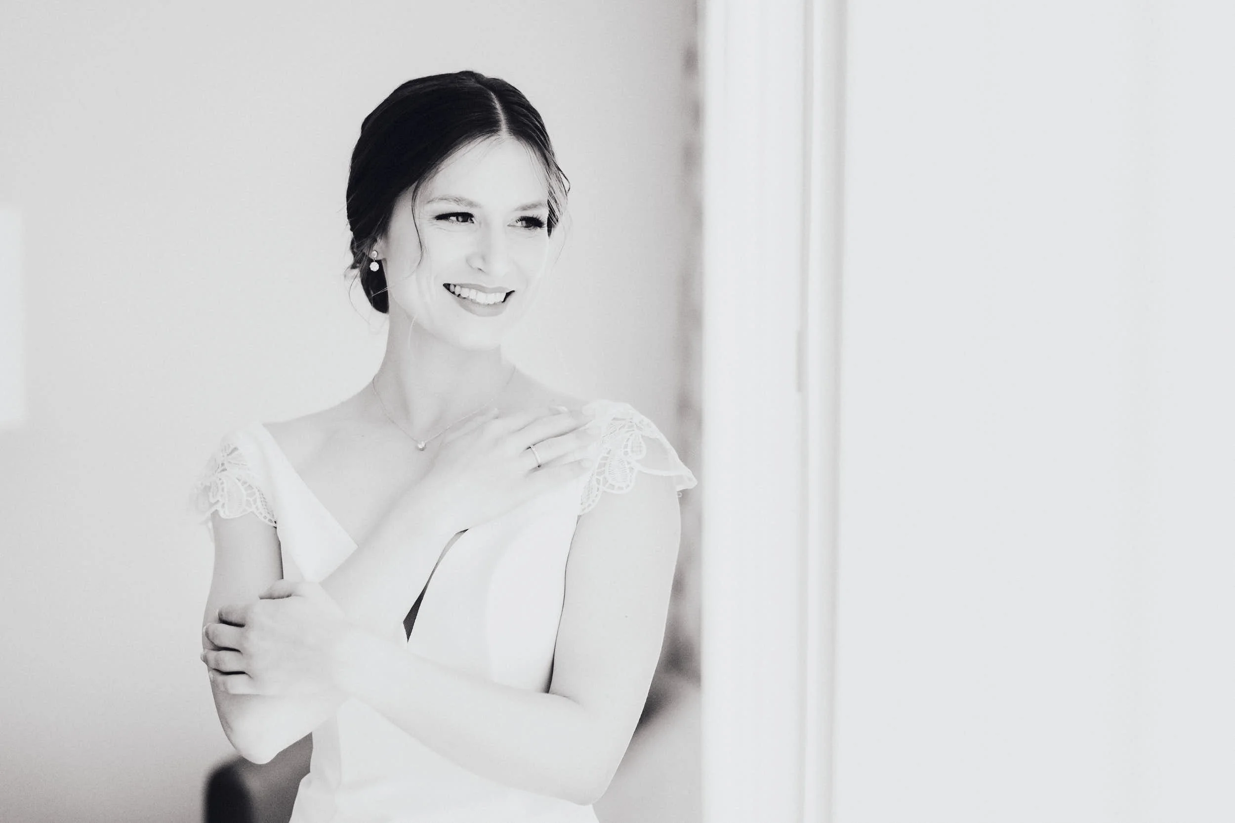 Schwarzweiß-Foto einer lächelnden Frau im weißen Kleid, die sich selbst umarmt. Sie steht in der Nähe eines Fensters.
