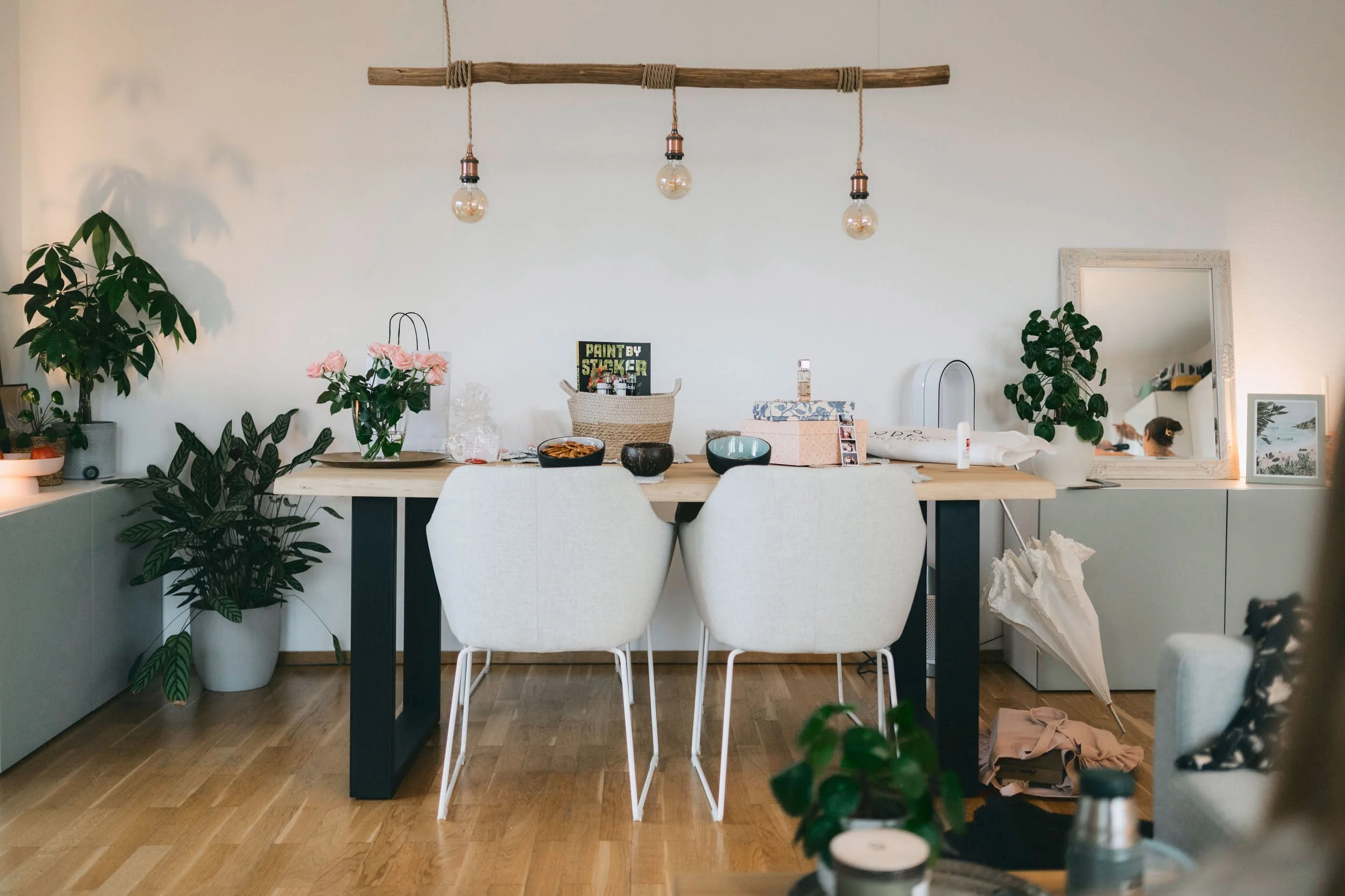 Ein Esstisch in einem Wohnzimmer mit weißen Stühlen, mehreren Pflanzen, einem Spiegel, Kerzen, Dekorationen und Schreibwaren. An der Decke hängt eine Leuchte mit drei Glühbirnen.