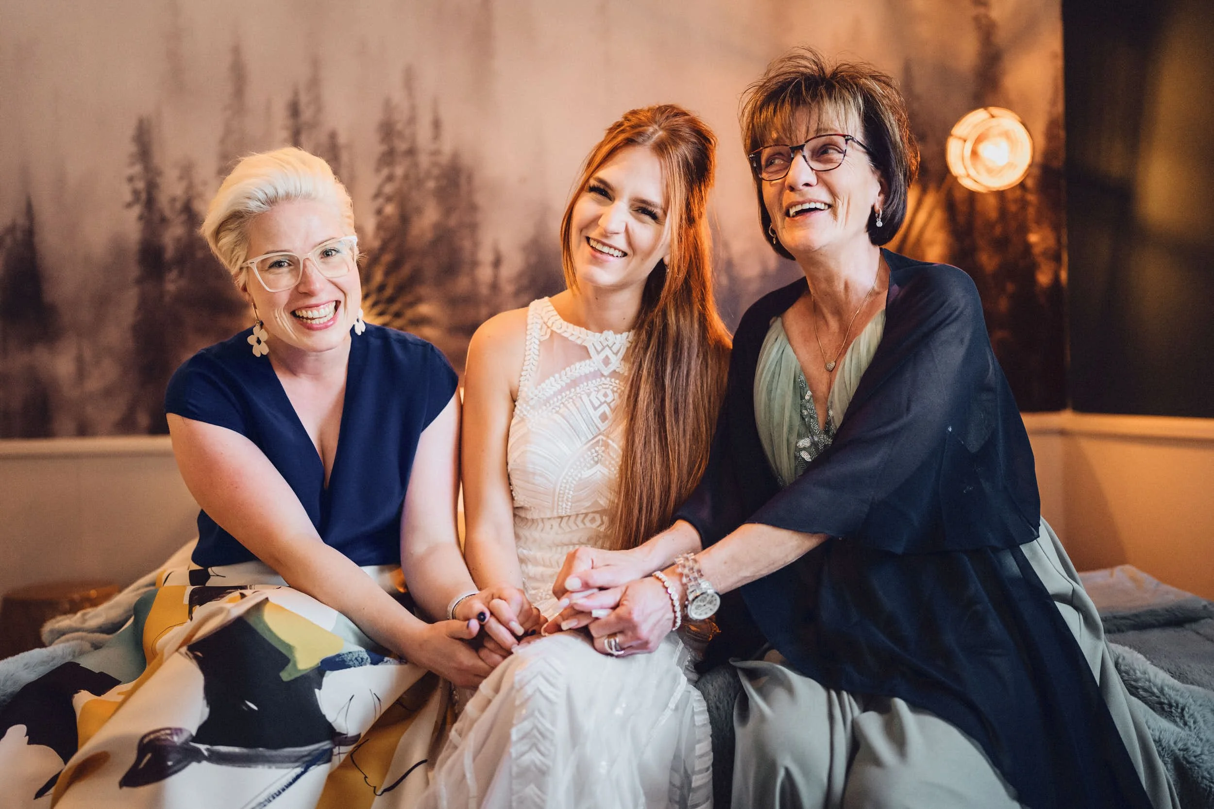 Three women sitting together, smiling, wearing elegant dresses, with a forest-themed backdrop.