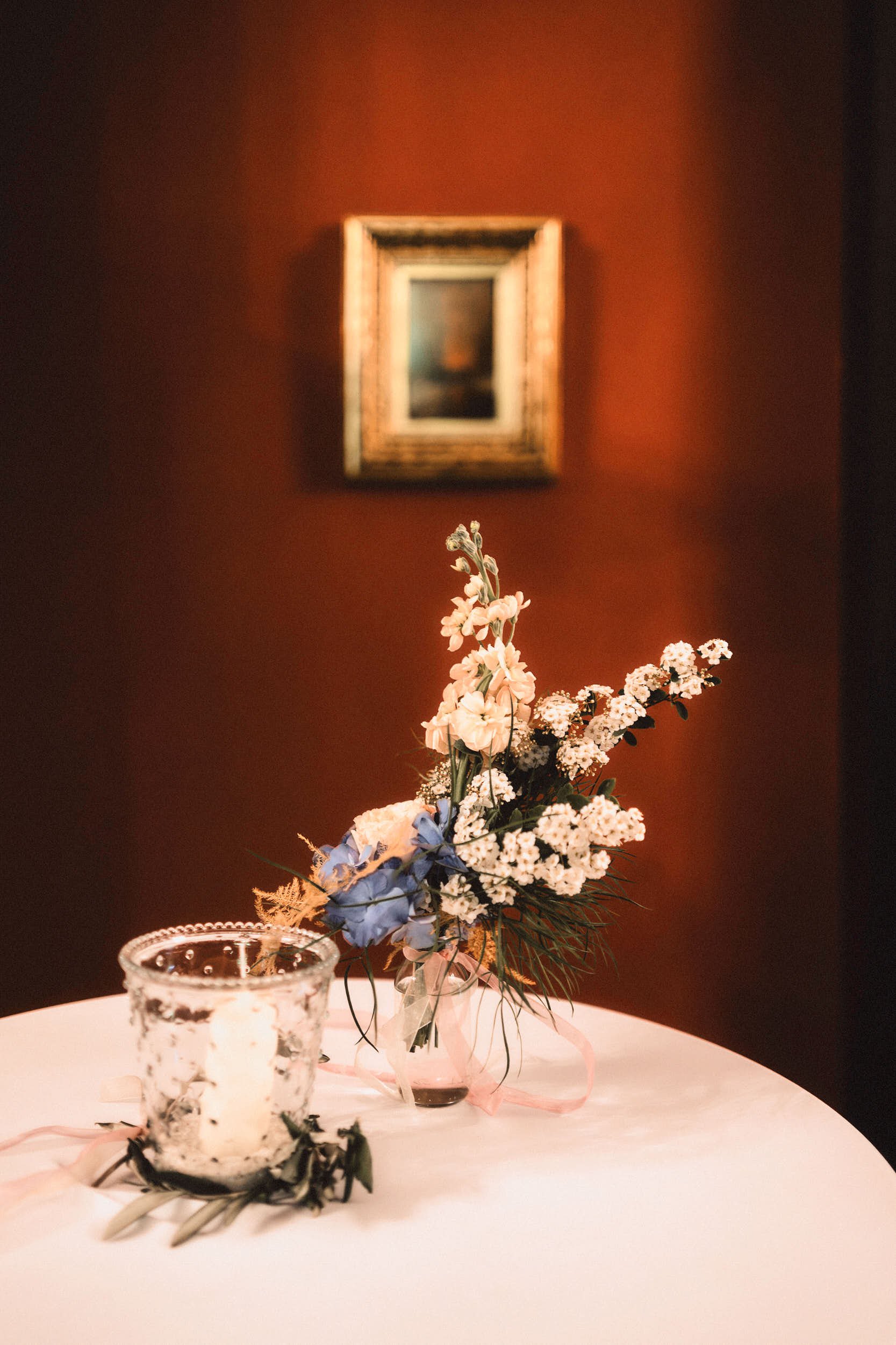 Blumen in einer Vase auf einem weißen Tisch, eine Kerze in einem Glasbehälter, an einer roten Wand mit einem kleinen, unscharfen Gemälde