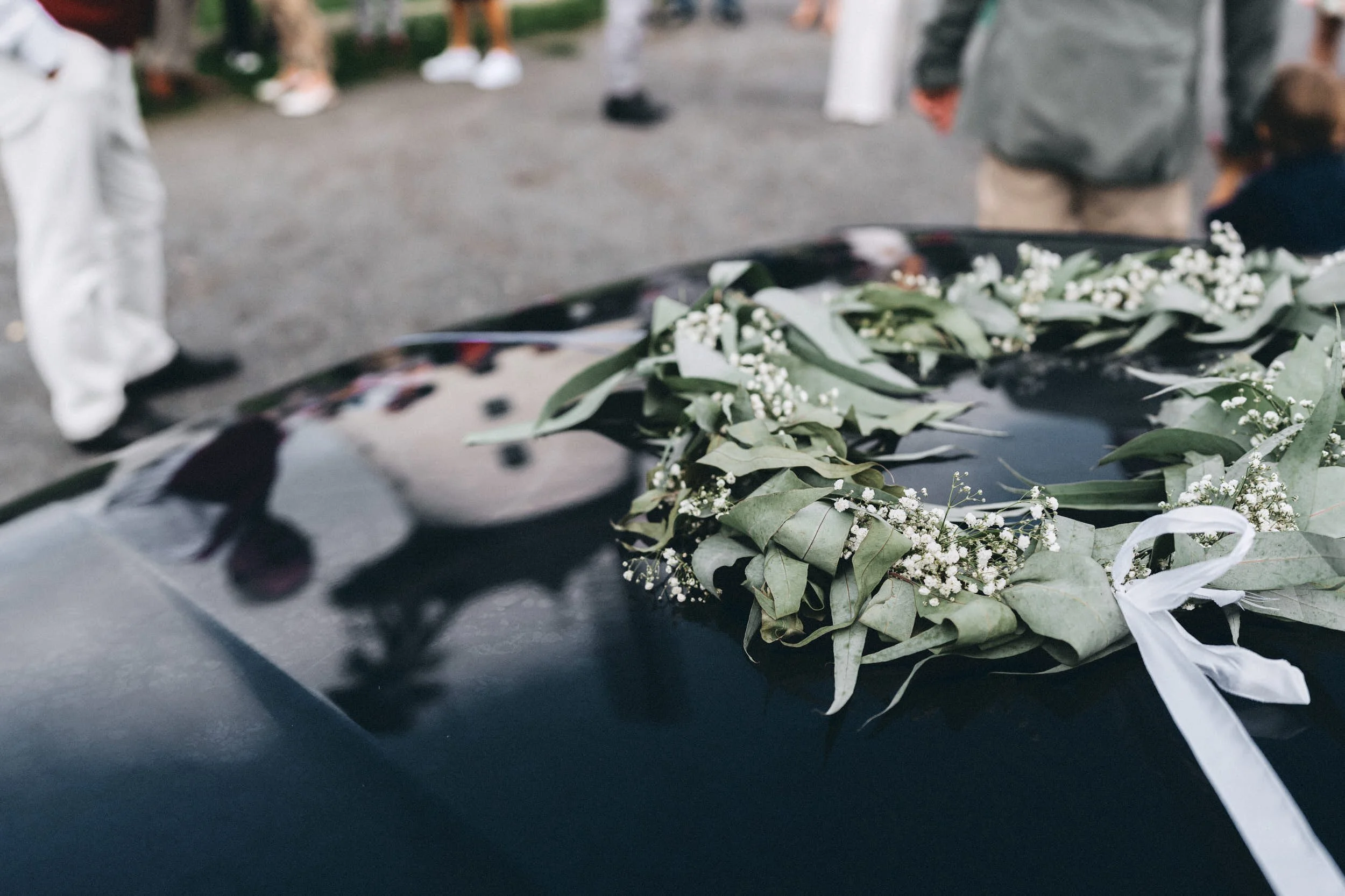 Blumenkranz mit weißen Blüten und grünen Blättern auf einem schwarzen Tisch, im Hintergrund Menschen bei einer Veranstaltung.